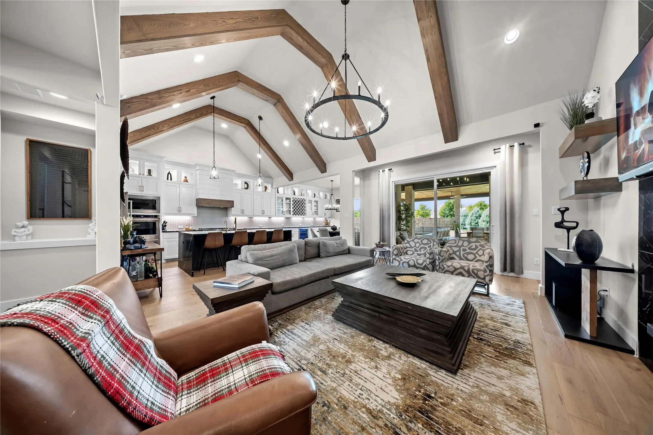 Vaulted ceilings with exposed wood beams, wood-finish flooring, a kitchen with white cabinetry and built-in appliances, and a sliding glass door to an outdoor living area