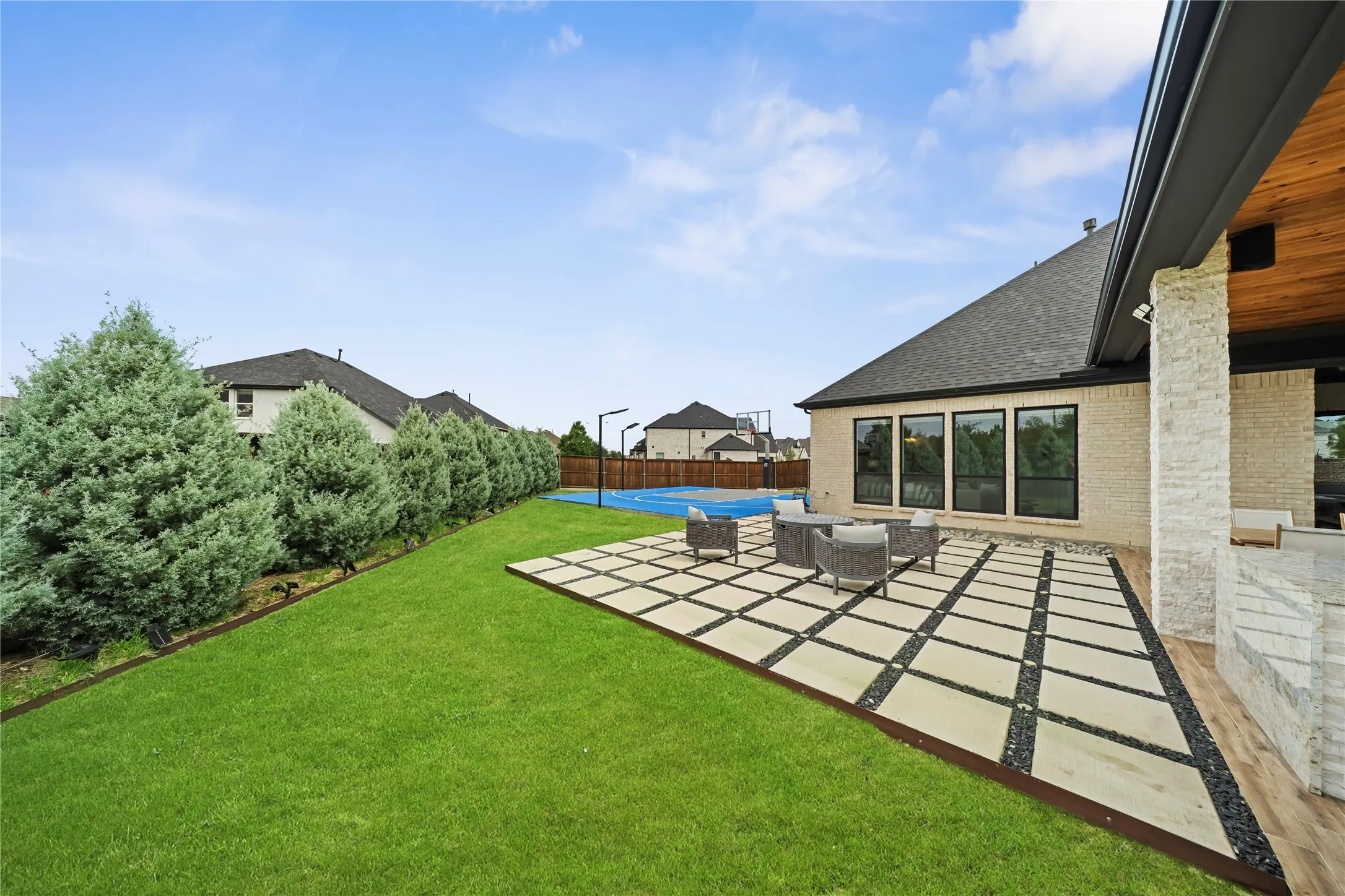 Expansive backyard featuring a manicured lawn, a half-court sports court, a stone-clad exterior, and a covered patio with a wood-finish ceiling