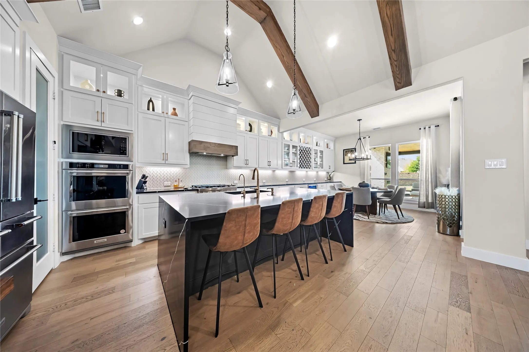 Vaulted ceiling with exposed wood beams, wood-finish flooring, custom white cabinetry, stainless steel double oven, and a large kitchen island with seating