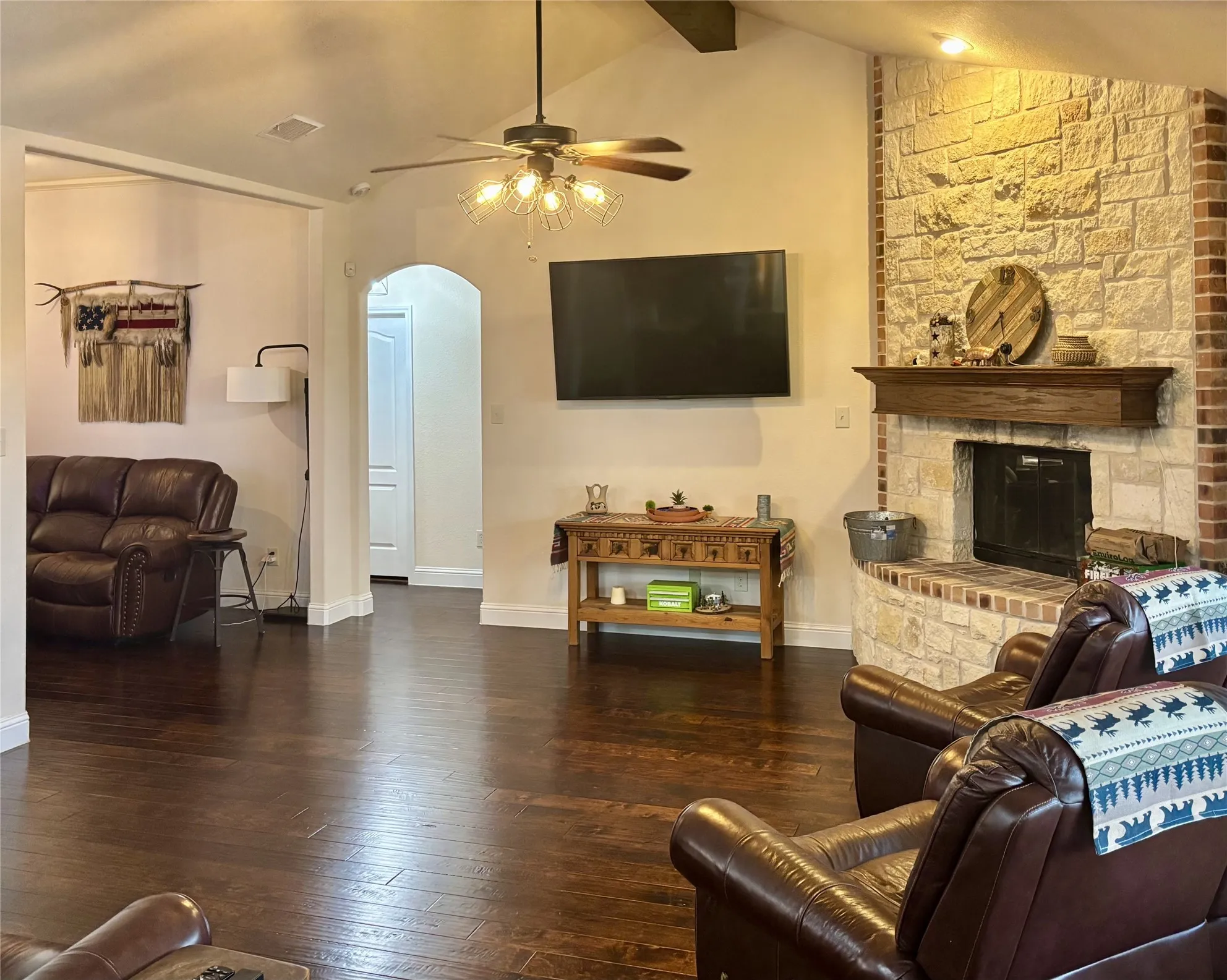 Spacious living area showcasing dark hardwood flooring, a wood-burning fireplace with a decorative brick surround, and a ceiling fan with integrated lighting