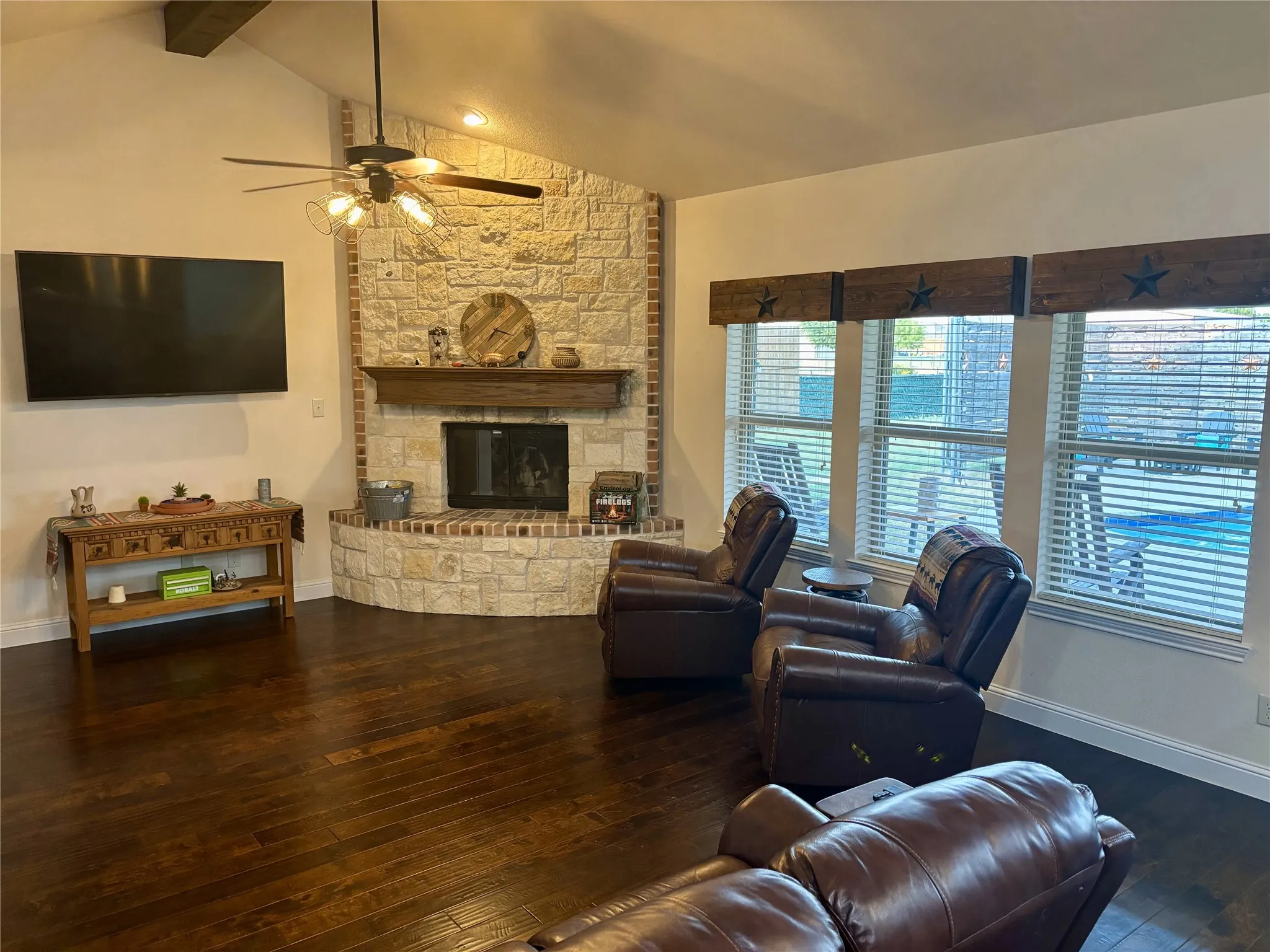 Vaulted ceiling with wood beam accent, stone-clad fireplace with wood mantel, dark wood-finish flooring, and multiple windows with blinds