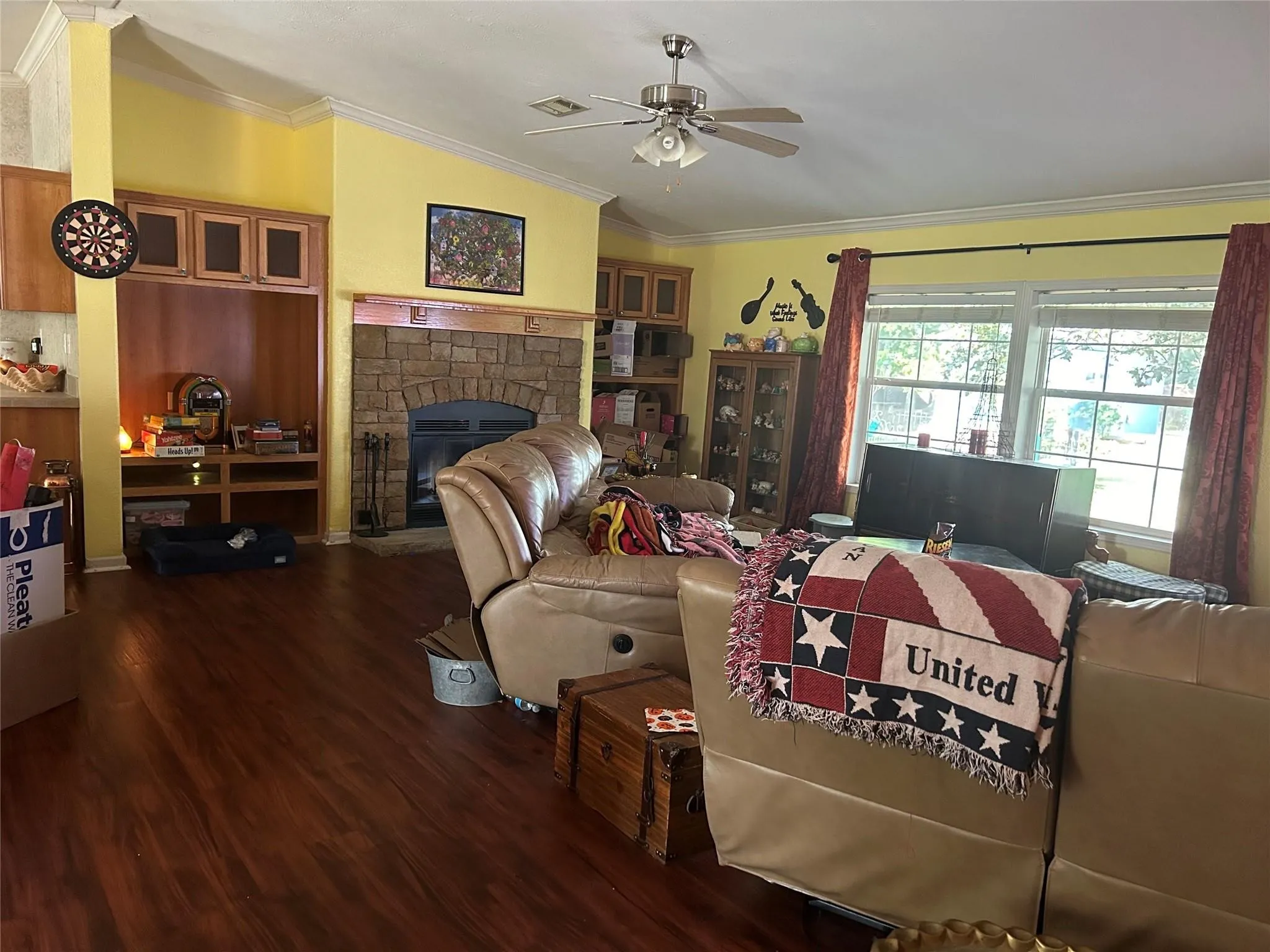 Wood-finish flooring throughout, stone-clad fireplace with hearth, built-in media cabinetry, ceiling fan, and crown molding details