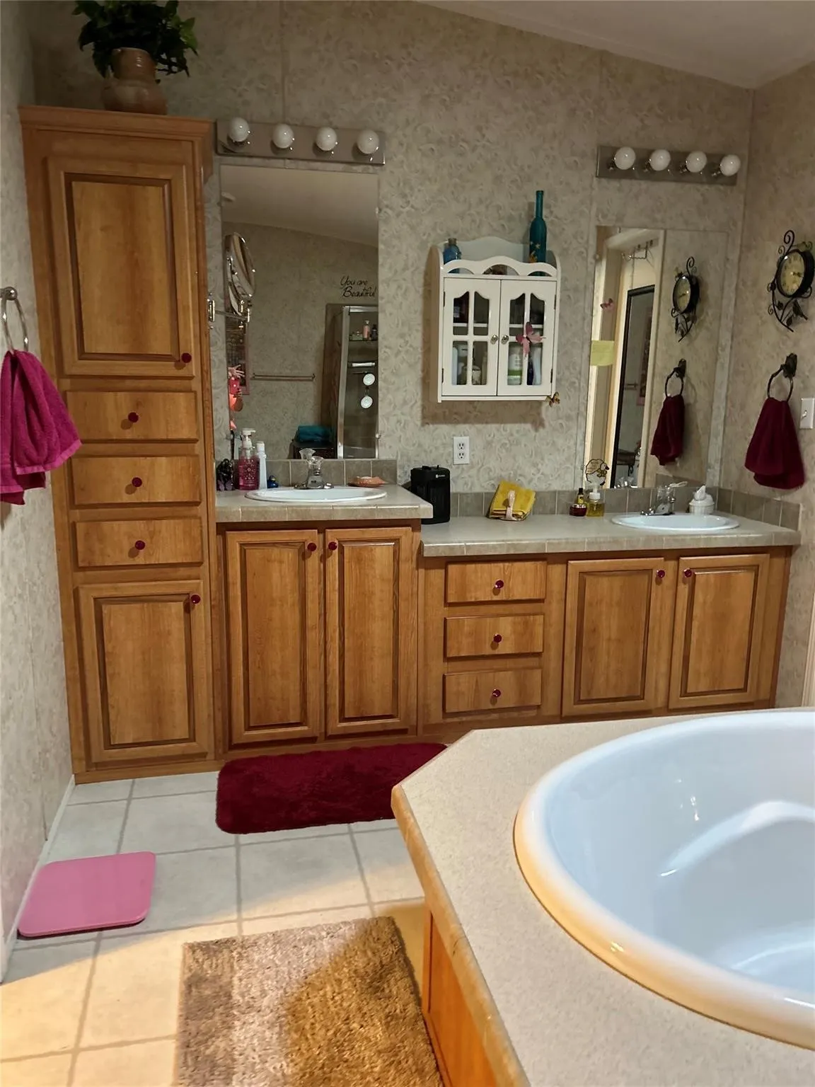 Bathroom featuring a dual vanity, light wood-finish cabinetry, built-in soaking tub, tile flooring, and wall-mounted mirrors with vanity lighting