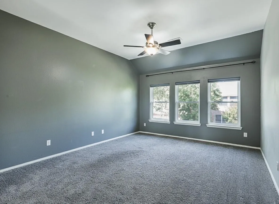 Bright corner bedroom showcasing neutral gray carpeting, ceiling fan with integrated lighting, and triple-pane windows flooding the space with natural light
