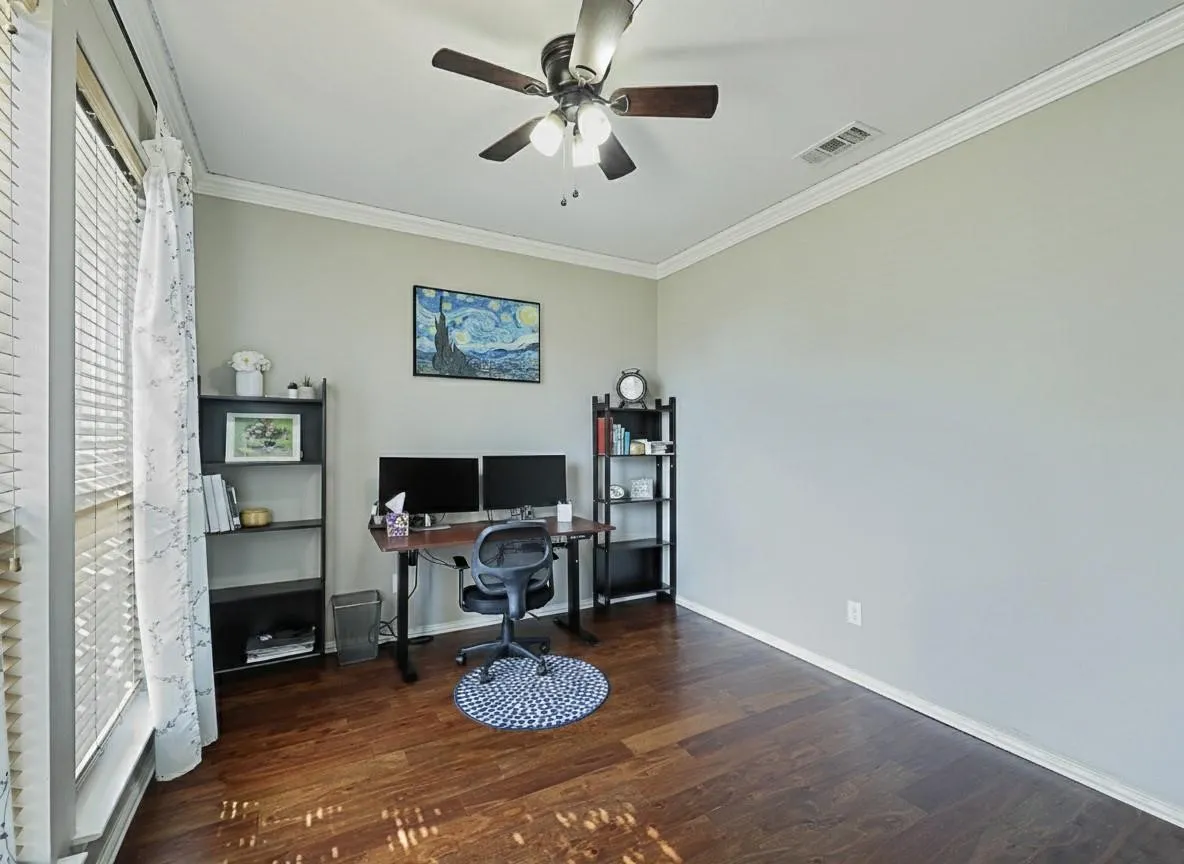 Dedicated home office featuring hardwood flooring, a built-in desk workspace with dual monitor setup, ceiling fan with integrated lighting, and abundant natural light from large windows with plantation shutters