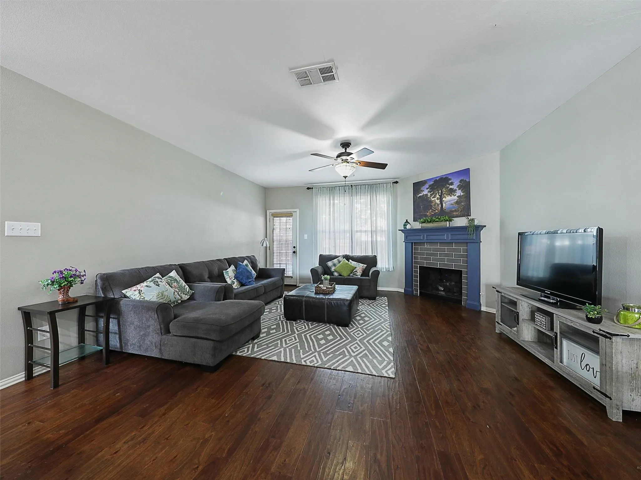 Spacious living room featuring rich wood-finish flooring, a fireplace with blue surround, ceiling fan, and large windows with sheer drapery
