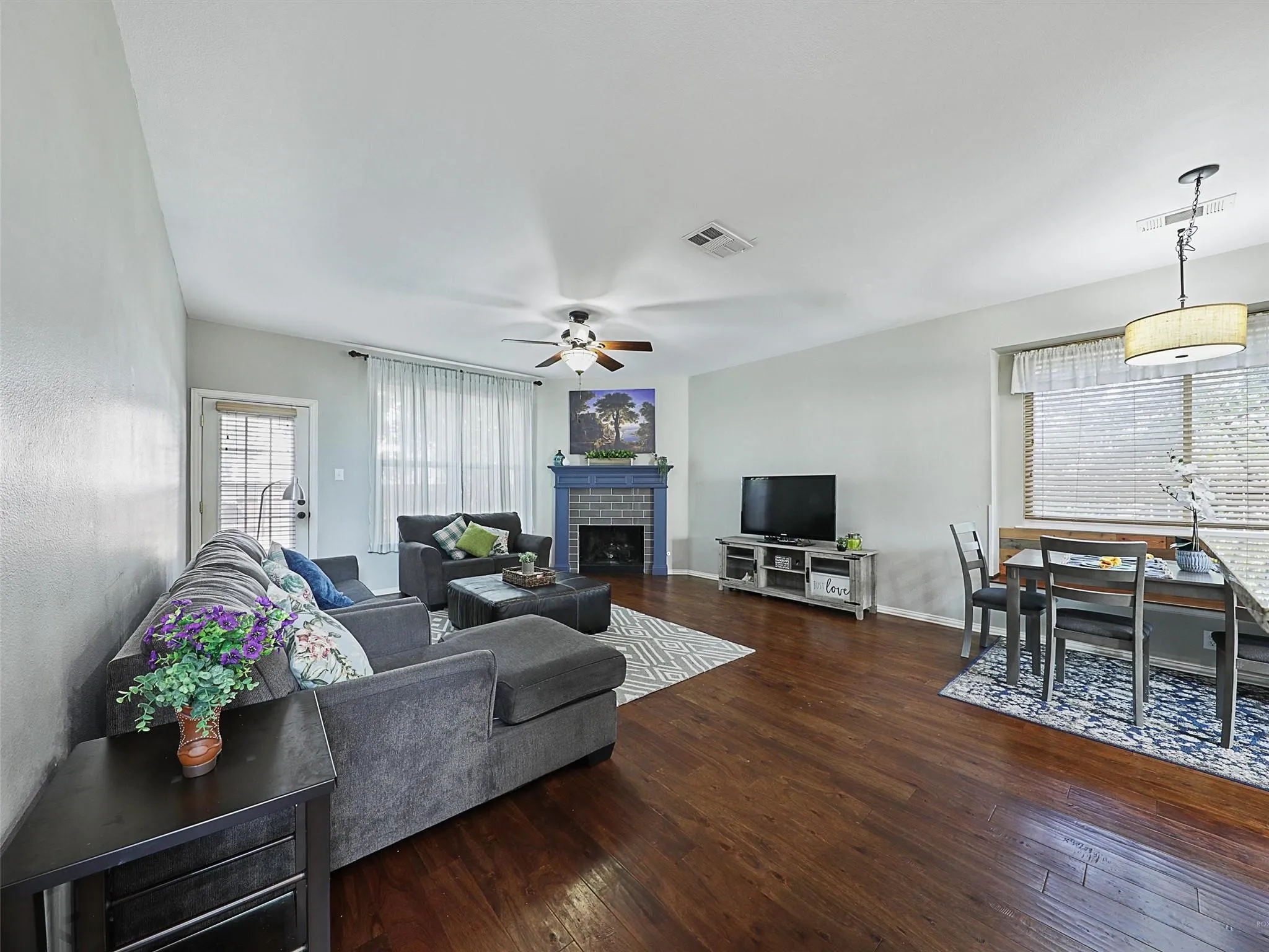 Open-concept living space featuring rich wood-finish flooring, a gray upholstered sectional, gas fireplace with surround, ceiling fan with integrated lighting, and abundant natural light from multiple windows with horizontal blinds