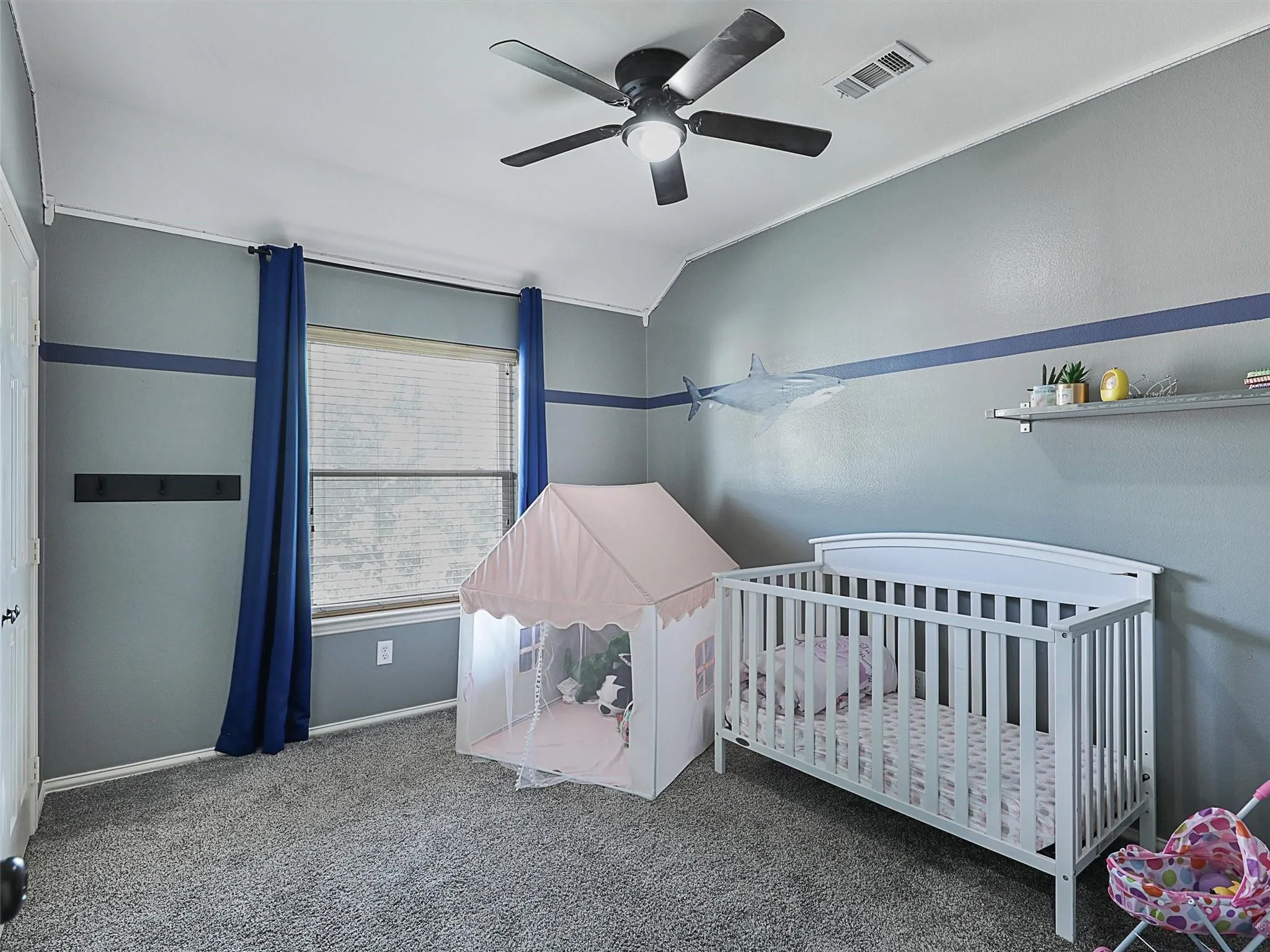 Bright nursery featuring soft blue walls with coordinating blue accent stripe, ceiling fan with integrated lighting, white crib, floating shelving, carpeted flooring, and abundant natural light from large window with blue drapery
