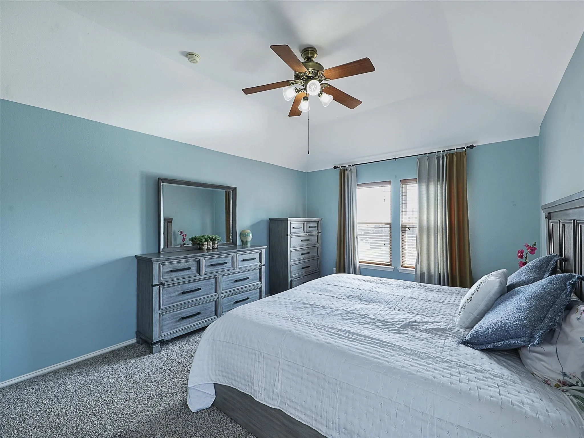 Serene primary bedroom showcasing soft blue walls, ceiling fan with integrated lighting, carpet flooring, and a wood-finish dresser with mirror