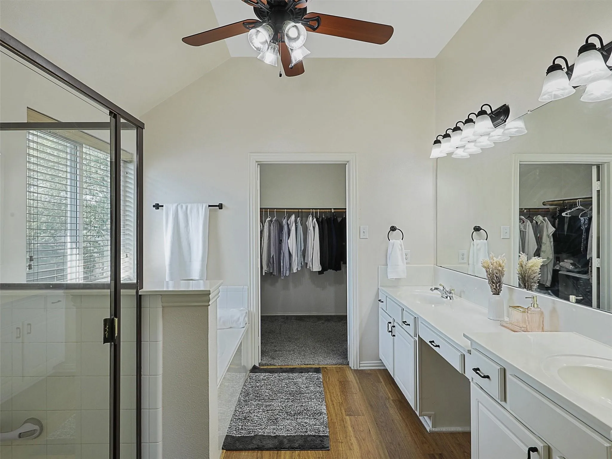 Vaulted ceilings with a ceiling fan, dual vanity with undermount sinks, wood-finish flooring, and a walk-in shower with a glass enclosure