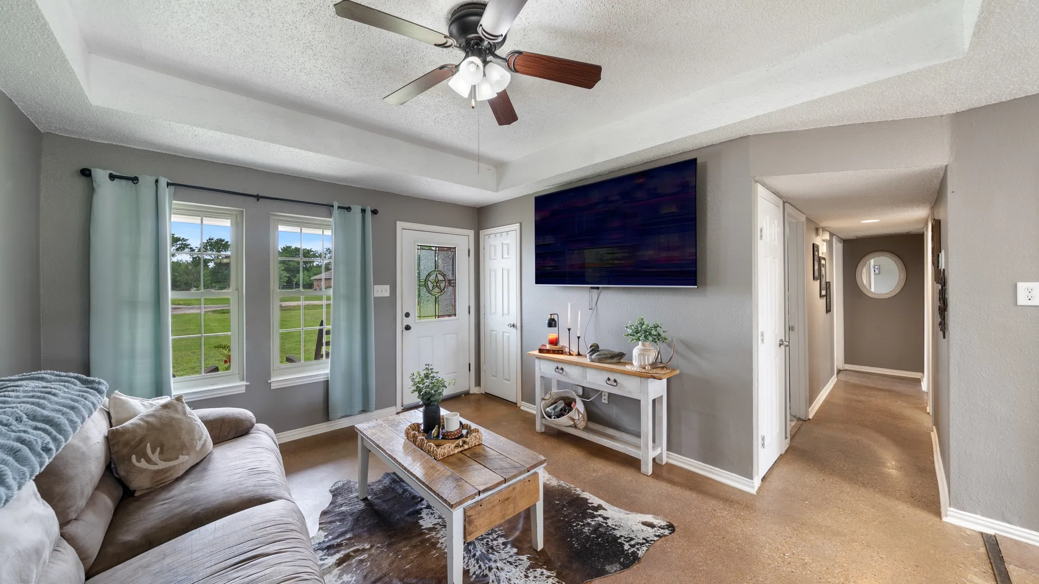 Bright, open living area featuring large glass doors with natural light, ceiling fan with integrated lighting, wall-mounted flat-screen television above built-in cabinetry with shelving, wood-finish flooring, and access to hallway and additional rooms