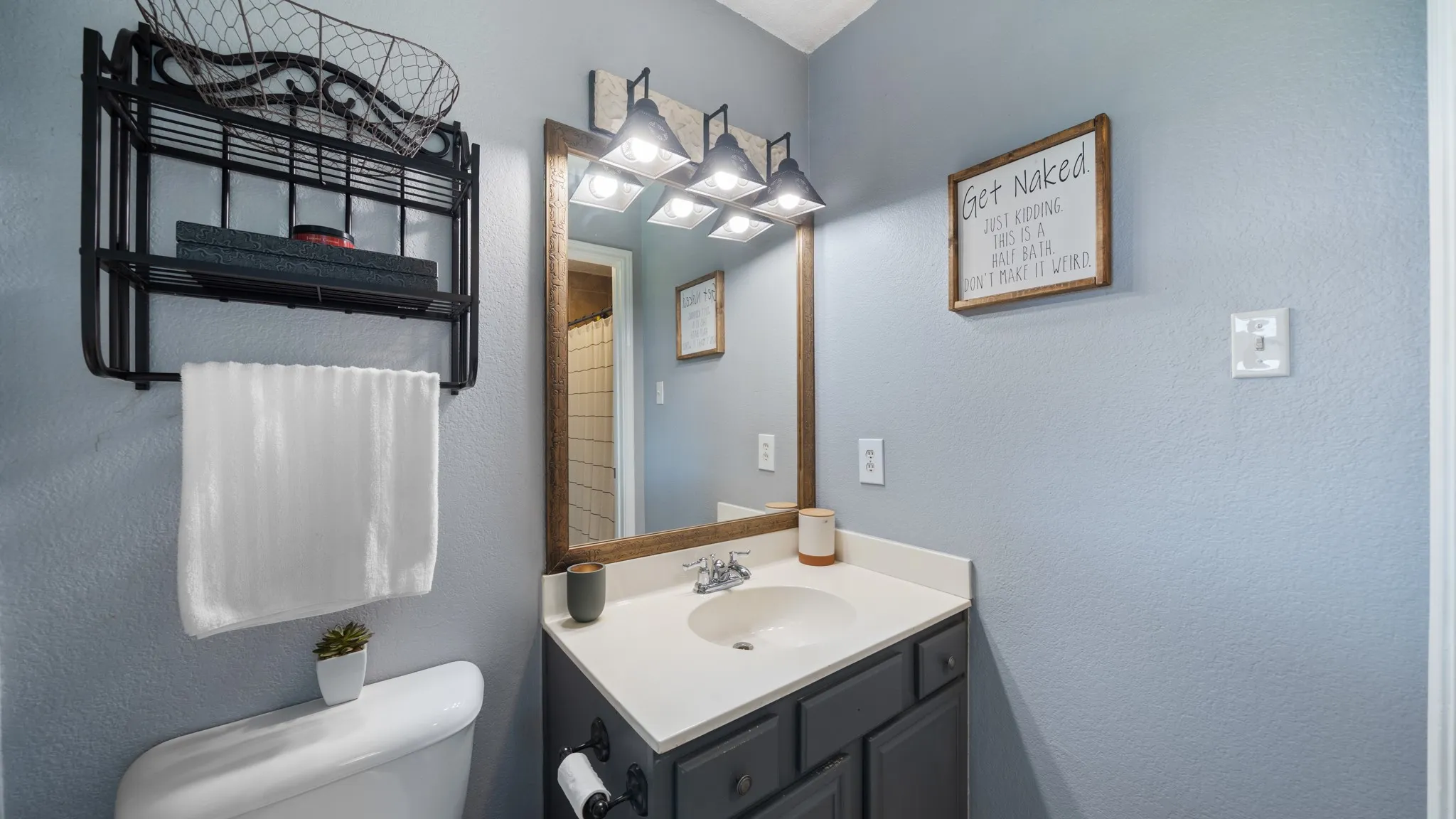 Refreshed bathroom featuring soft blue walls, a dark vanity with white countertop and undermount sink, large framed mirror with integrated lighting, wall-mounted shelving with decorative storage, and updated fixtures throughout