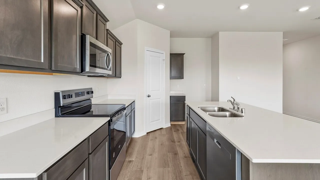 Modern galley kitchen featuring dark cabinetry with brushed metal hardware, white quartz countertops, stainless steel appliances including a microwave and electric range, and wood-finish flooring throughout