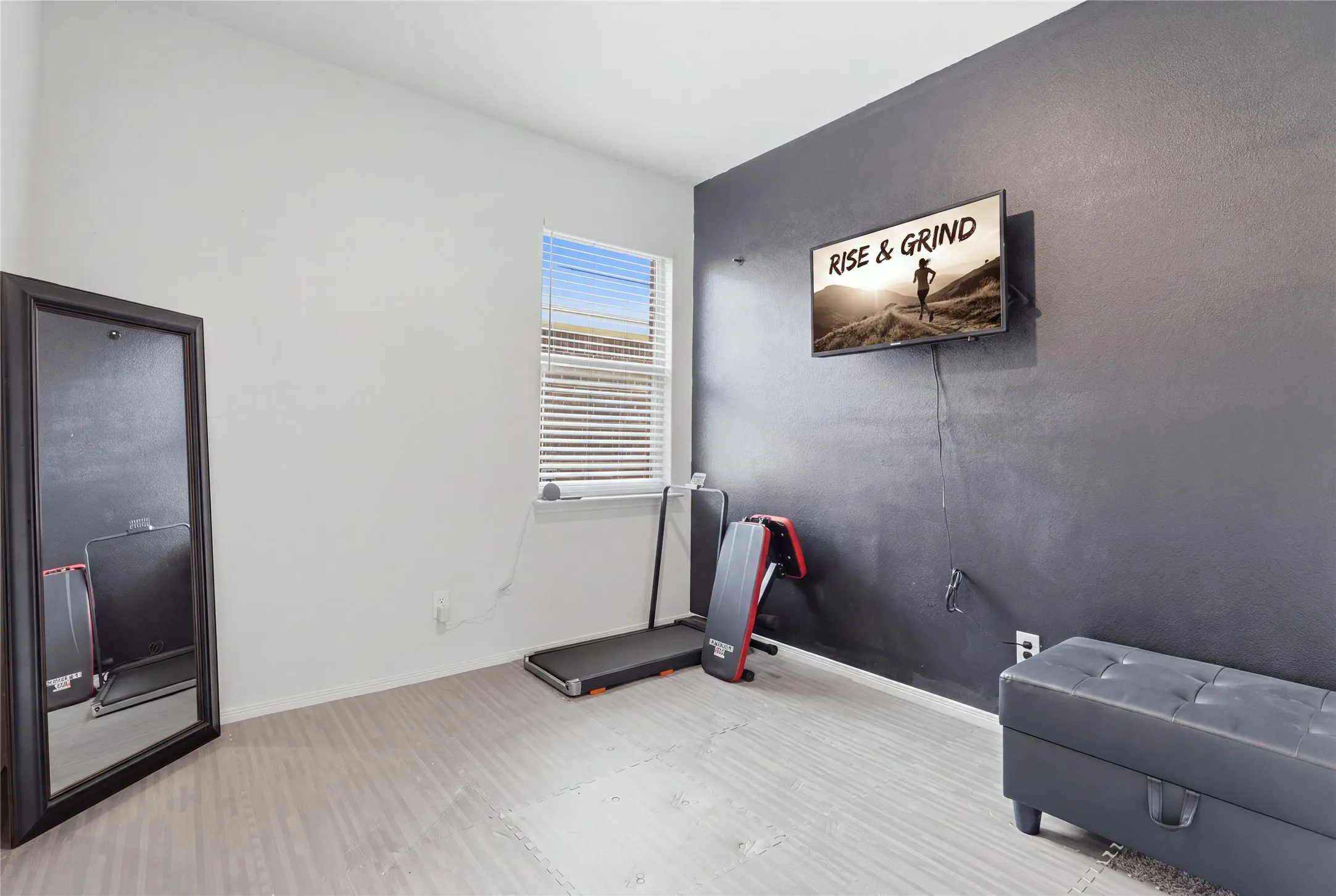 Versatile fitness room featuring light wood-finish flooring, large window with horizontal blinds, dark accent wall, wall-mounted television, and built-in storage cabinetry