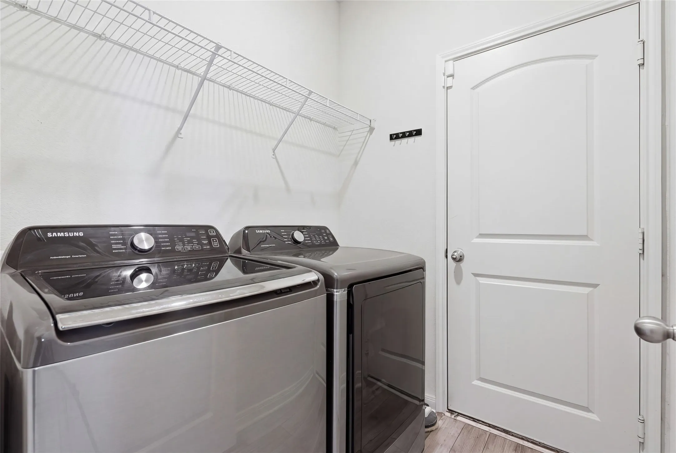 Dedicated laundry room featuring Samsung front-load washer and dryer with modern control panels, wire shelving for storage, wood-finish flooring, and a white arched-panel door for convenient access