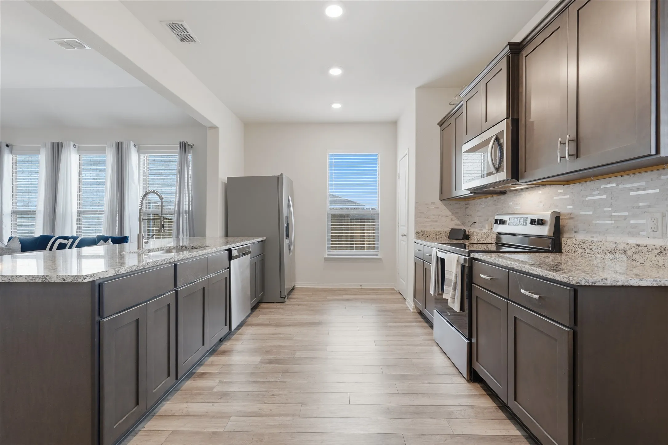 Open-concept kitchen with dark wood cabinetry, light granite countertops, stainless steel appliances, and a tile backsplash