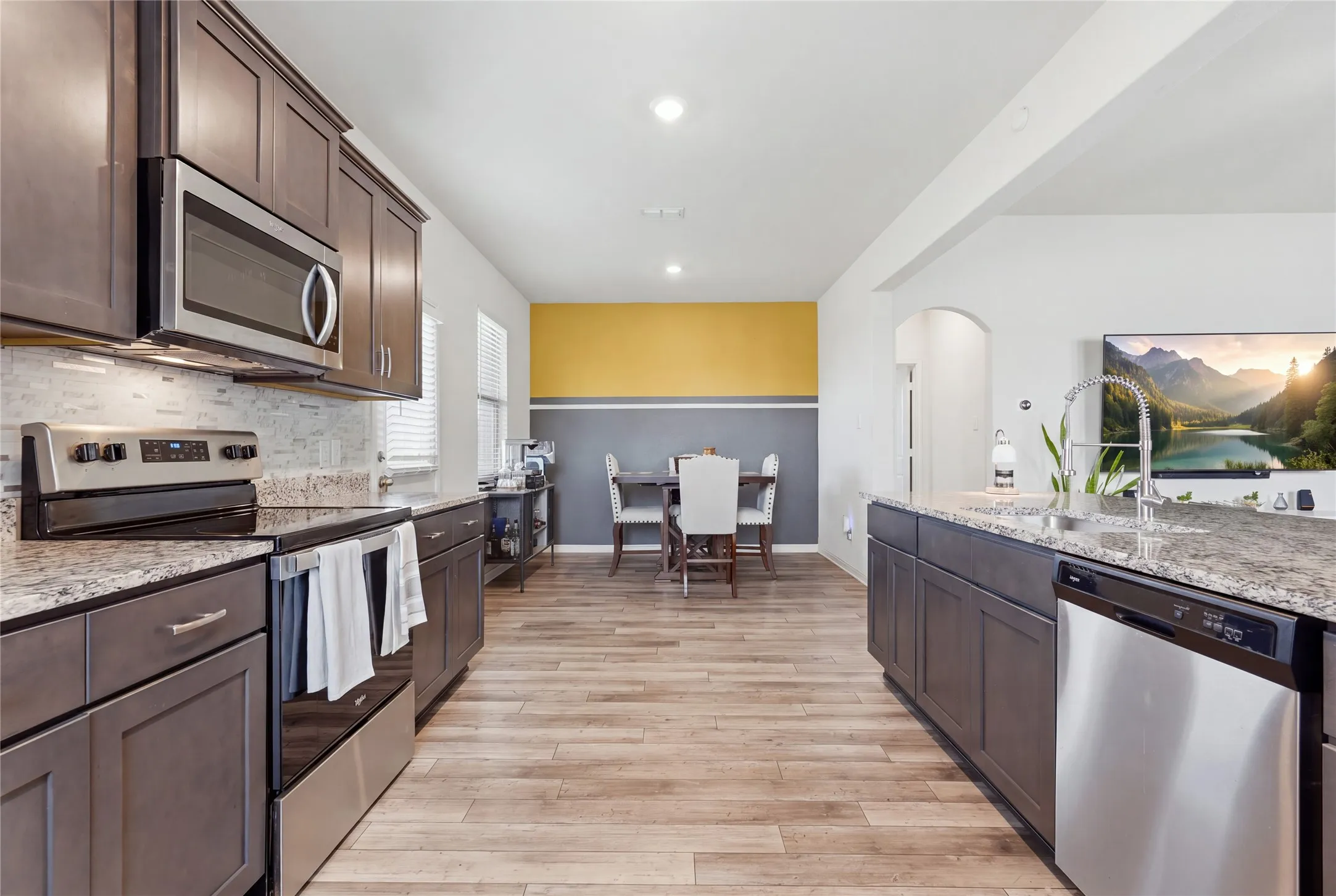 Modern kitchen and dining area featuring dark cabinetry with stainless steel appliances, granite countertops, subway tile backsplash, wood-finish flooring throughout, and a two-tone accent wall with soft blue lower paneling and warm yellow upper section