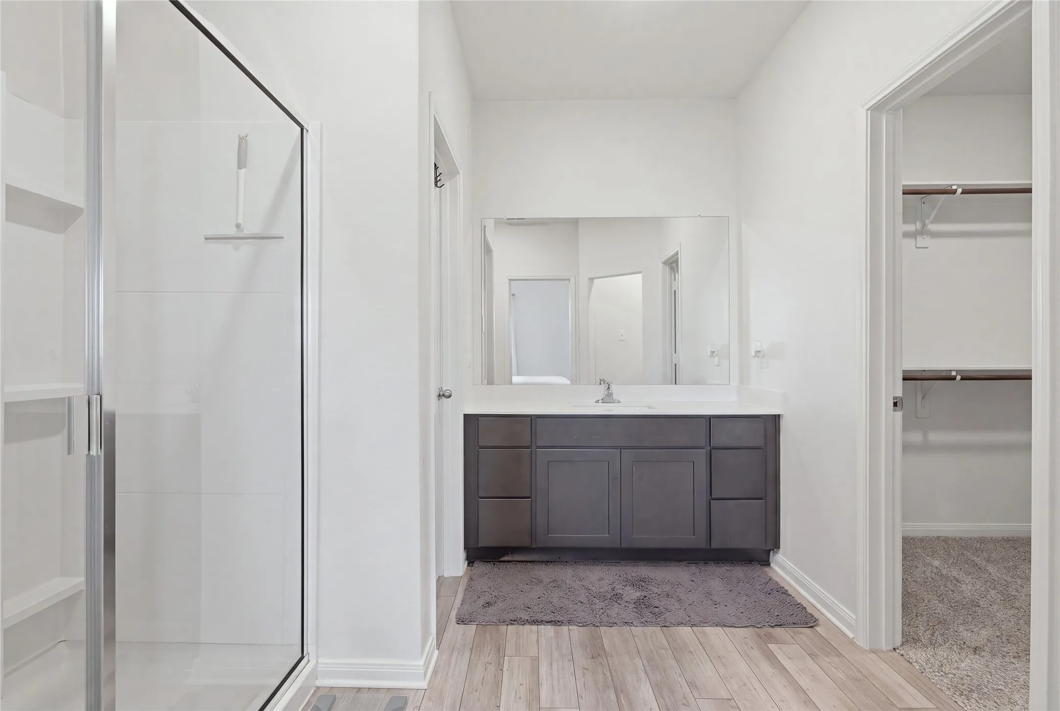 Full glass-enclosed shower with built-in shelving, a single vanity with a gray cabinet, a white countertop, and wood-finish flooring