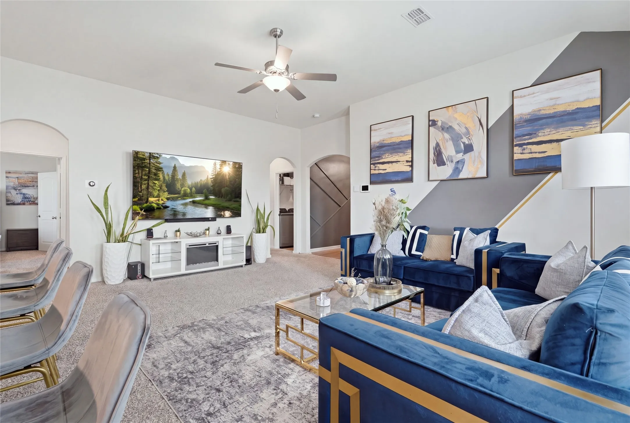 Open-concept living area featuring a white built-in media console with integrated storage, ceiling fan with integrated lighting, neutral wood-finish flooring, and a staircase with metal railings