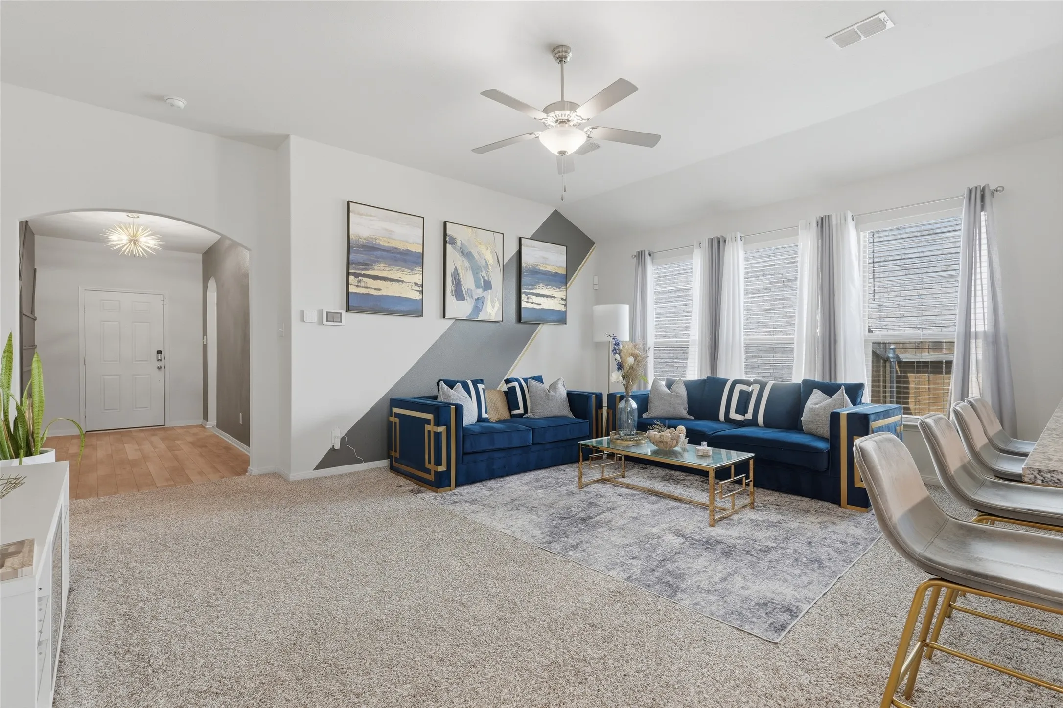 Bright, open living area showcasing neutral carpet flooring, multiple windows with plantation shutters, and a ceiling fan with integrated lighting