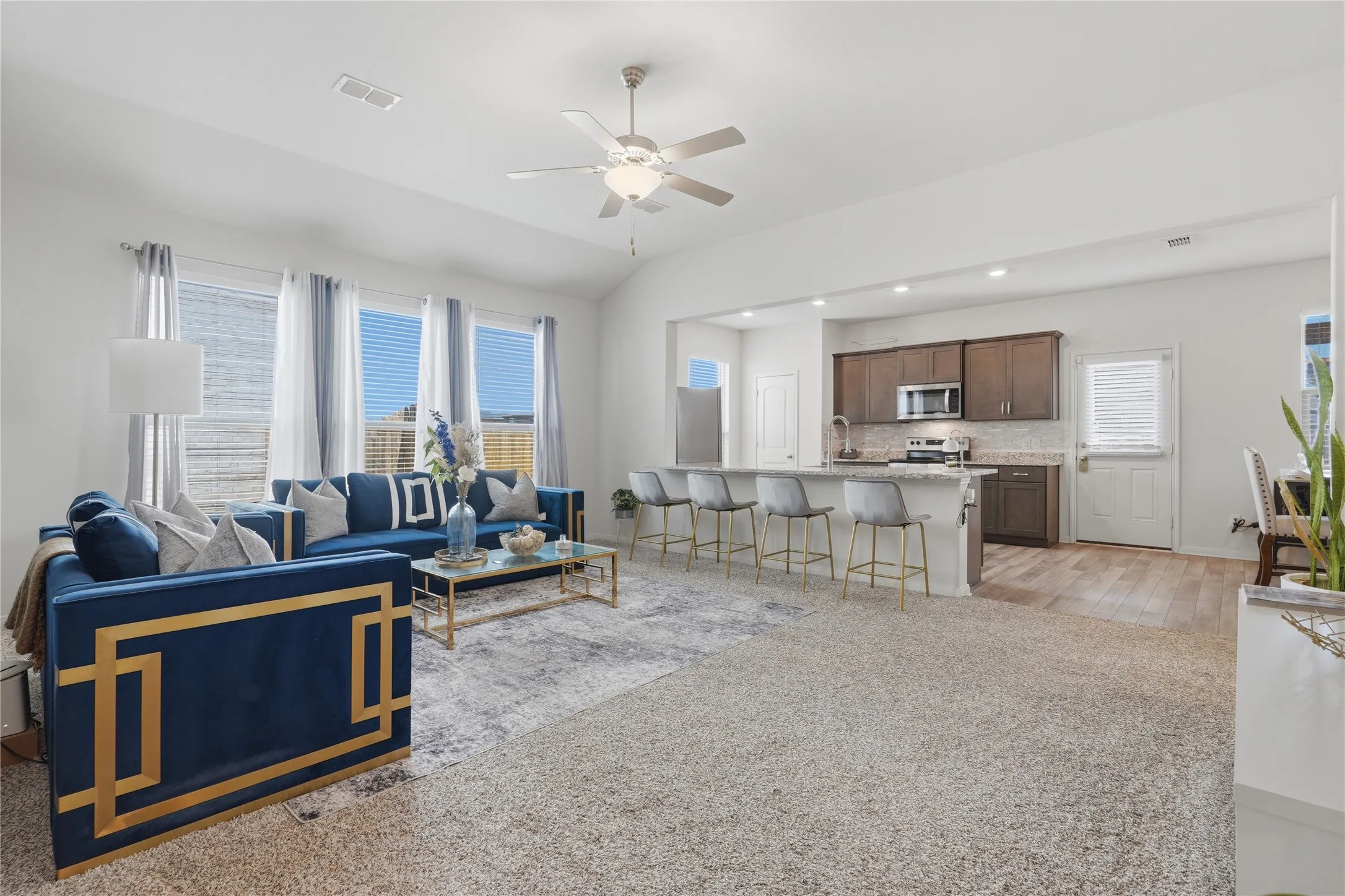 Open-concept living and kitchen area featuring a spacious floor plan with neutral carpeting, ceiling fan with integrated lighting, stainless steel kitchen appliances, wood-finish cabinetry, and large windows with natural light