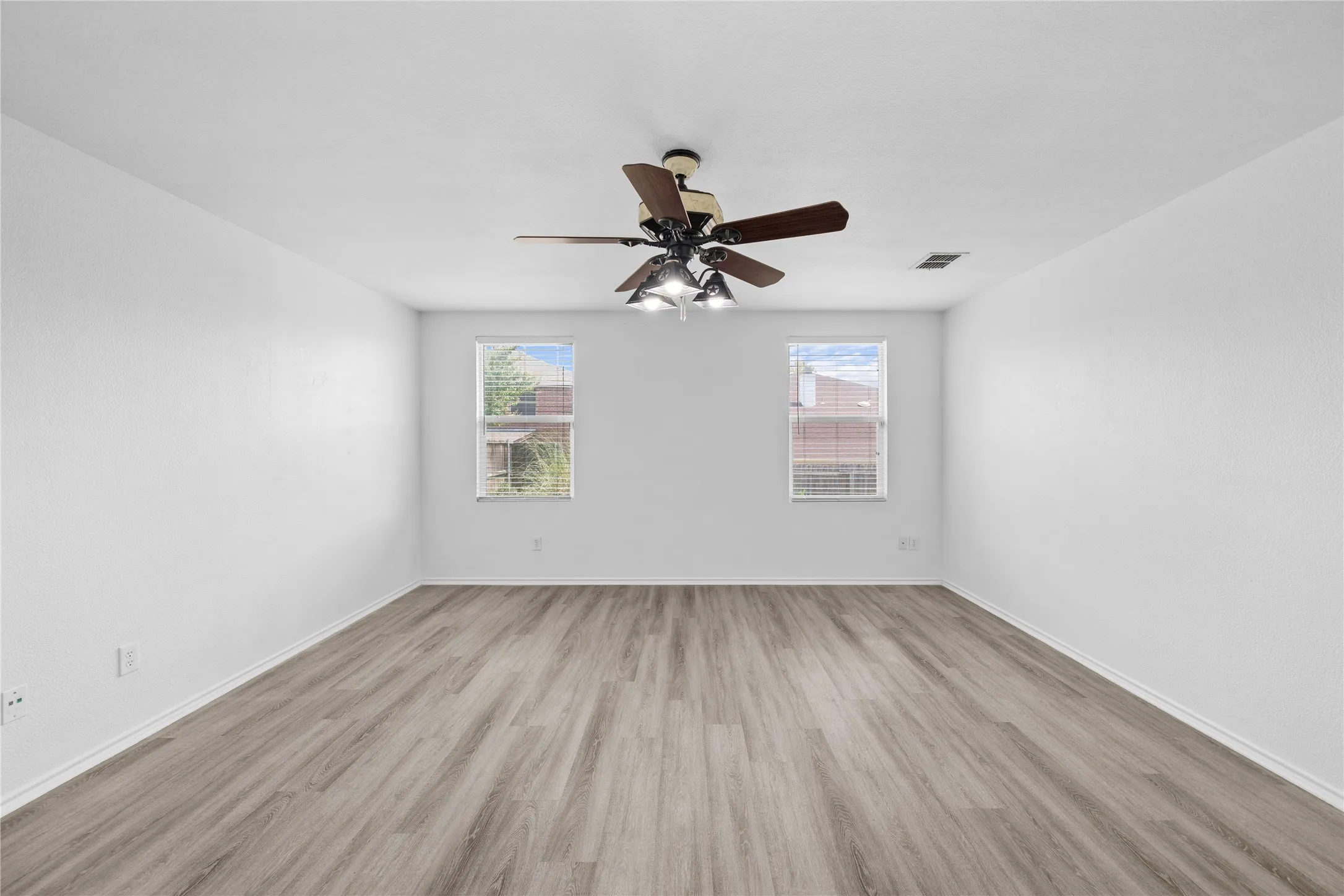 Spacious room featuring wood-finish flooring, white walls, two windows, and a ceiling fan with integrated lighting