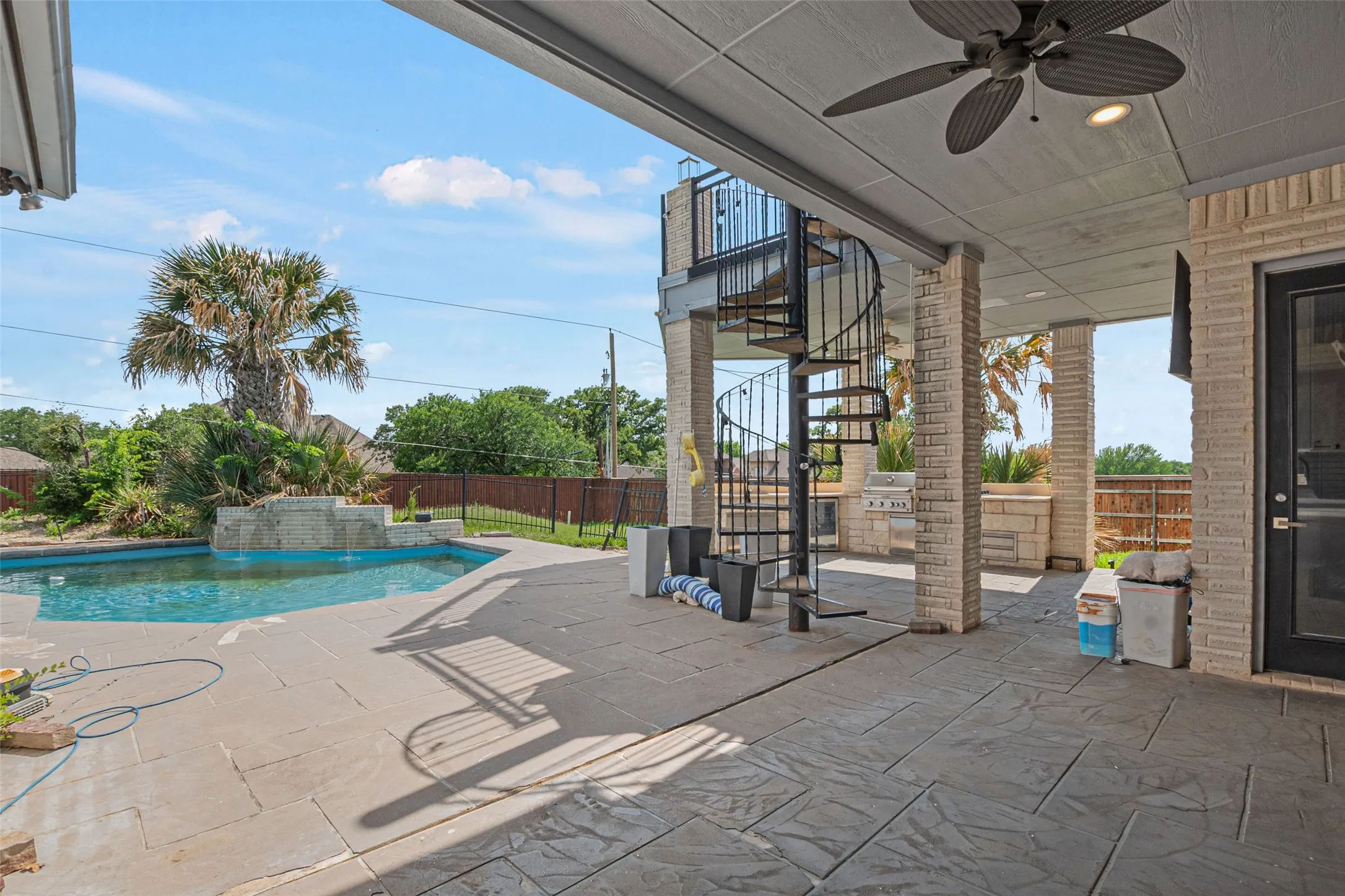 Expansive covered patio with ceiling fan and stacked-stone columns overlooking a resort-style saltwater pool, sun deck with decorative concrete pavers, and mature landscaping including palm trees
