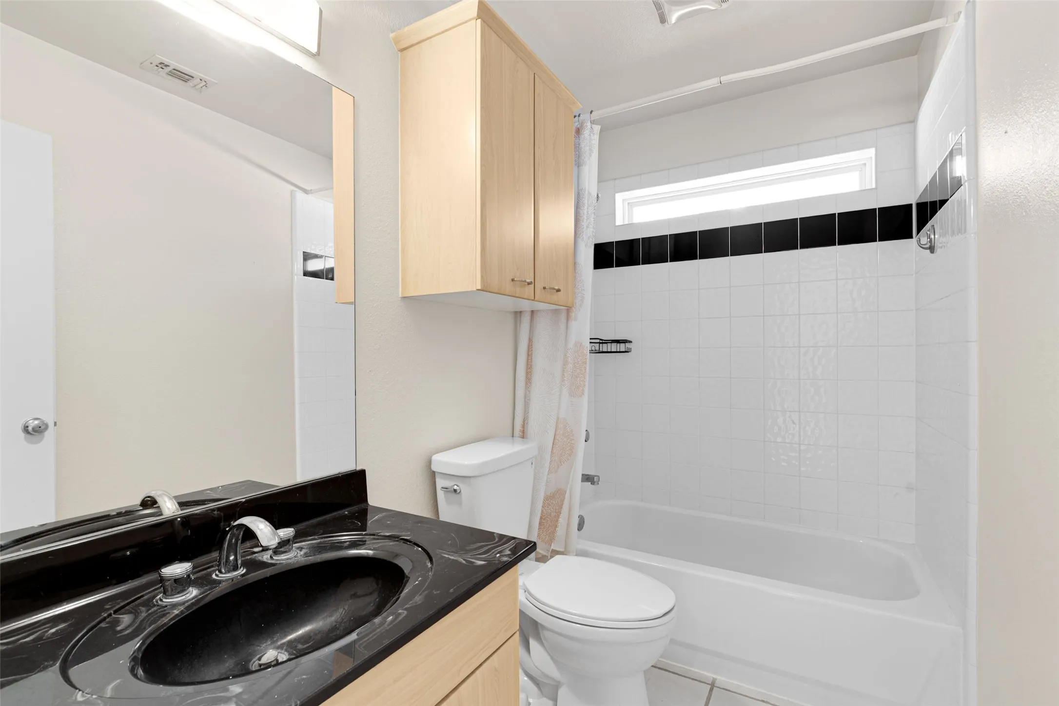Modern bathroom featuring a black granite-style countertop with an oval vessel sink, wood-finish cabinetry with ample storage, a white subway tile shower surround with contrasting black tile accent strip, and a separate toilet with a window for natural light