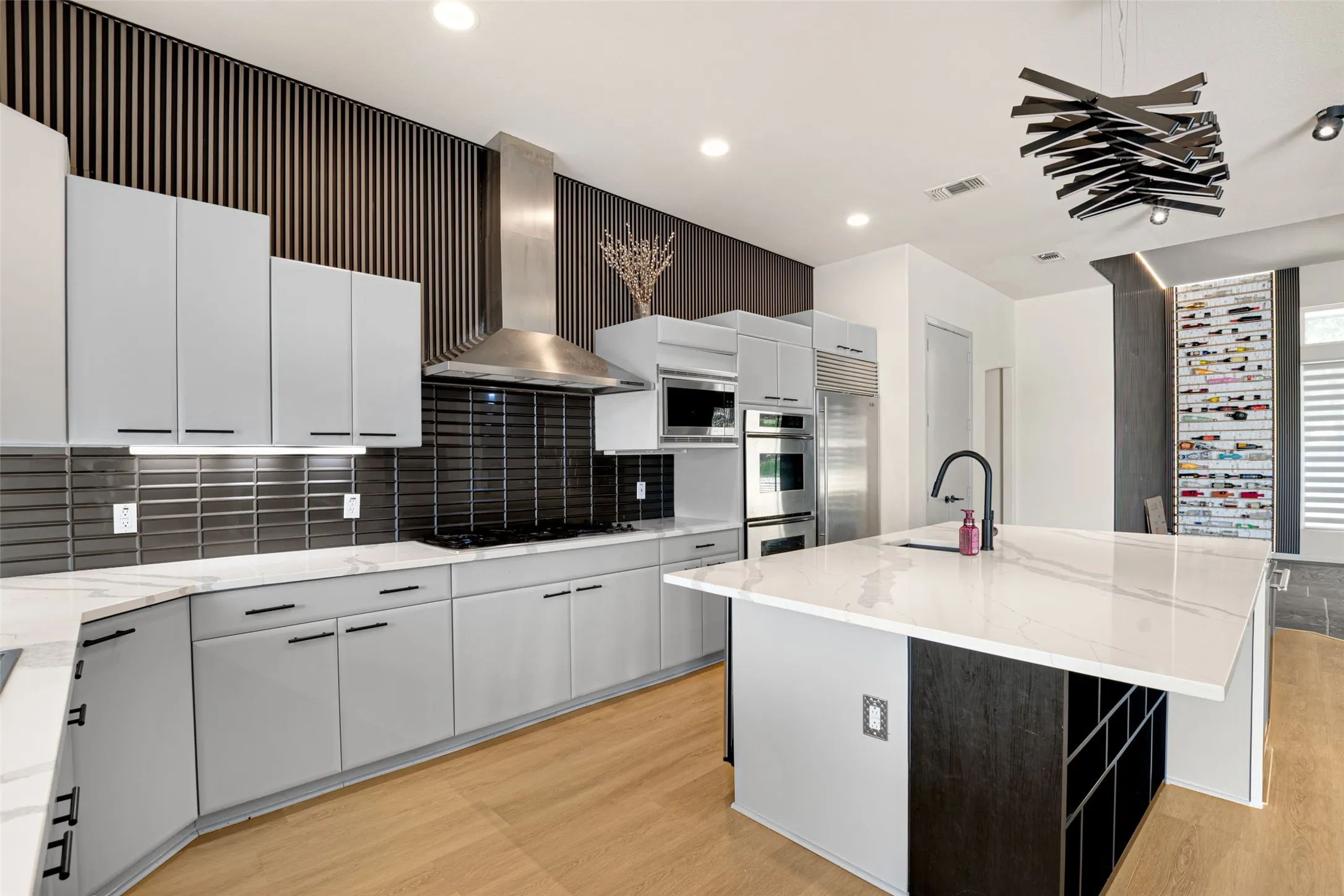 Modern kitchen featuring light gray cabinetry, a central island with a waterfall countertop, integrated stainless steel appliances, a unique decorative light fixture, and a built-in wine storage system