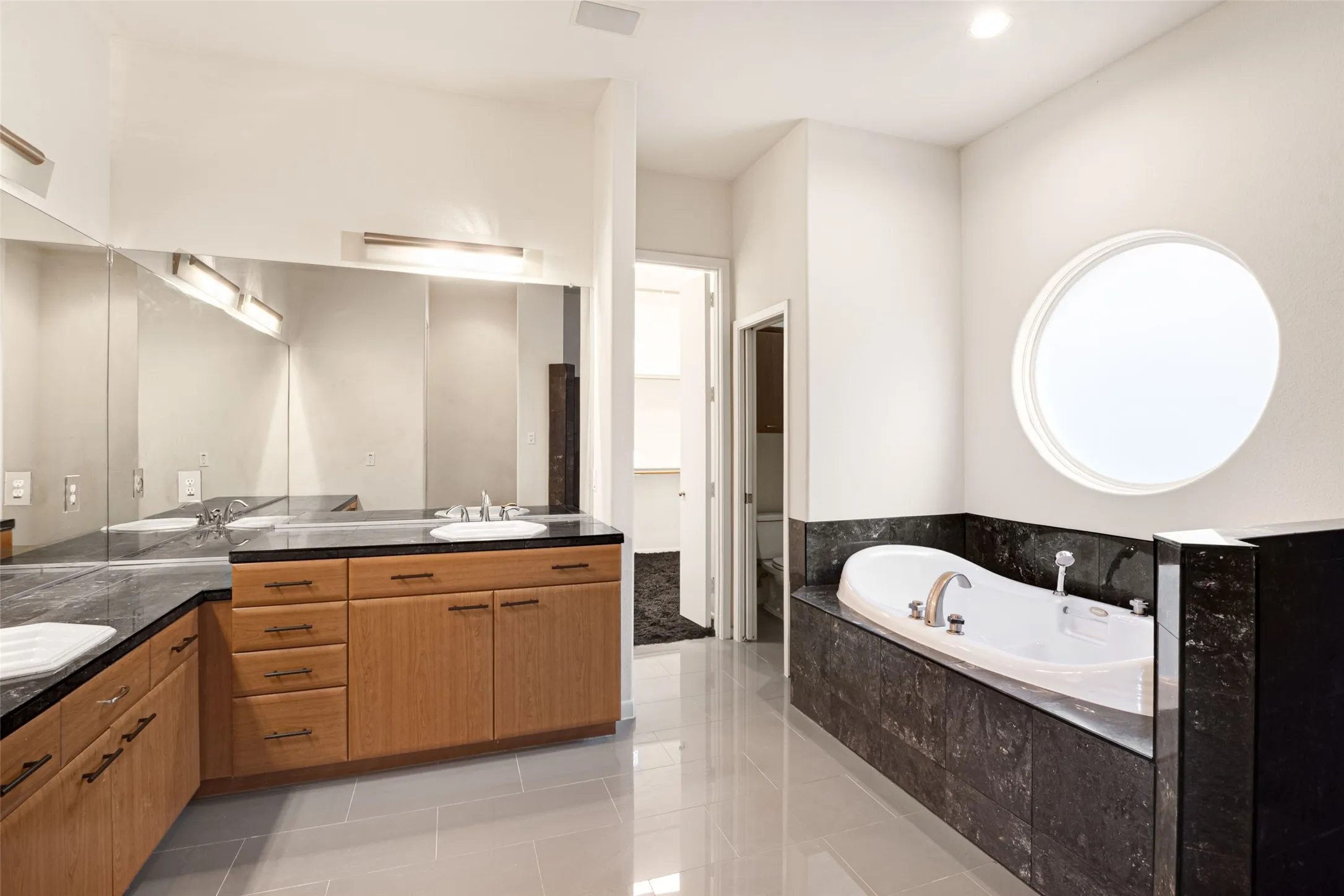 Spa-inspired primary bathroom featuring dual vanities with wood-finish cabinetry and dark stone countertops, a soaking tub set within dark tile surround, polished stone flooring throughout, large circular skylight providing natural illumination, and contemporary chrome fixtures
