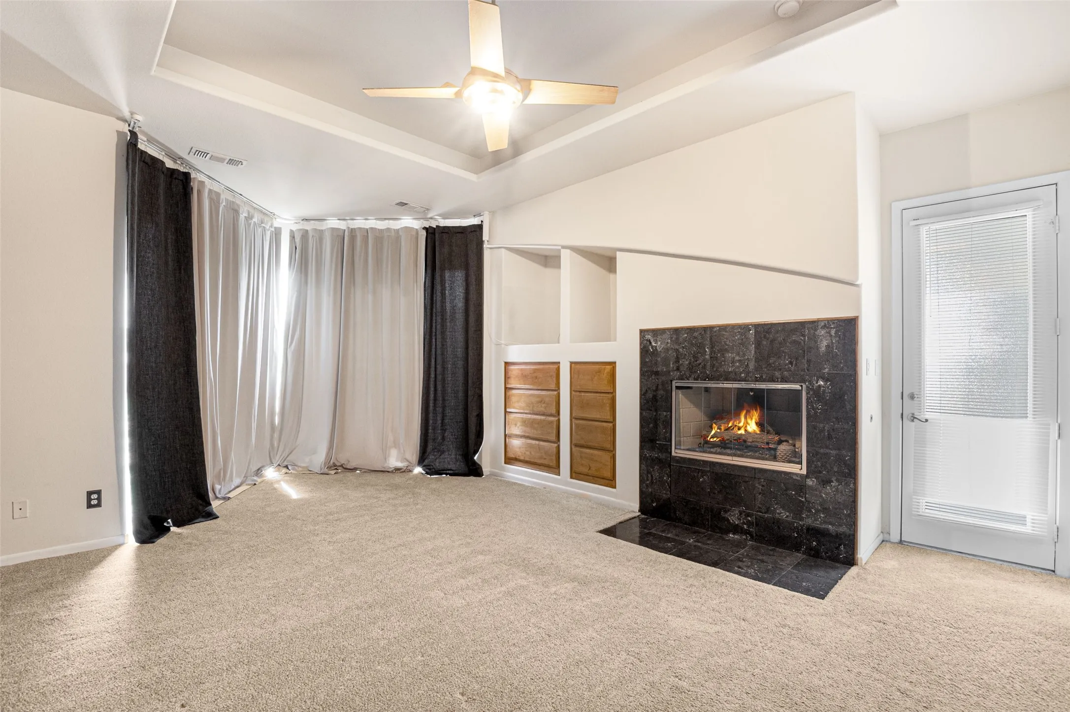 Spacious primary suite featuring neutral carpeting, a gas fireplace with dark stone surround and wood-finish cabinetry, recessed ceiling lighting with a ceiling fan, floor-to-ceiling windows with layered drapery, and a glass-paneled exterior door providing natural light
