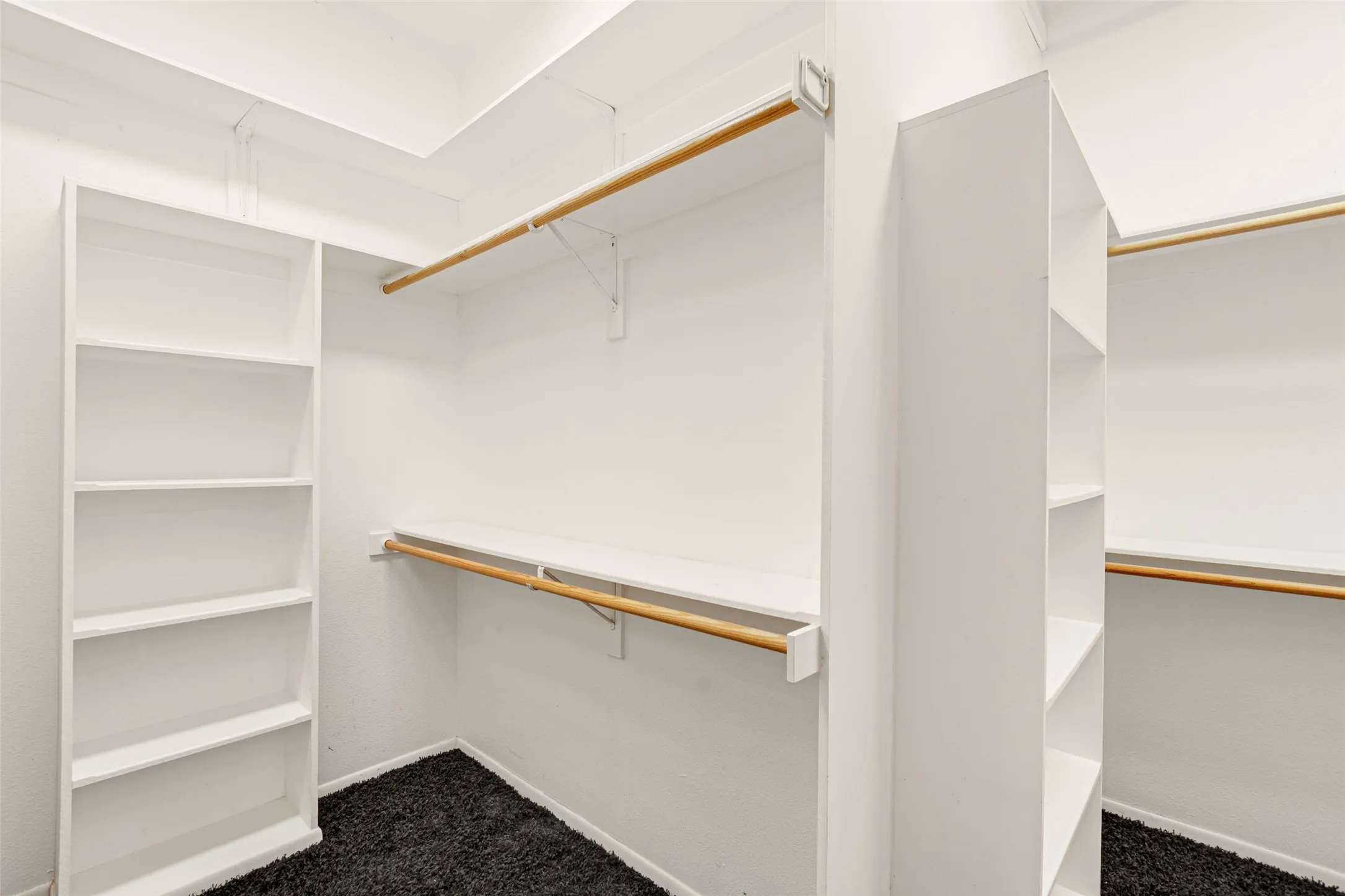 Spacious walk-in closet featuring custom built-in shelving and storage cubbies along one wall, dual wooden hanging rods mounted at varying heights, and a dark-finish flooring base
