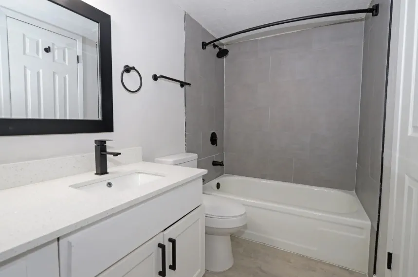 Modern bathroom featuring a white vanity with integrated sink and matte black hardware, gray tile shower surround with frameless glass enclosure, white subway tile backsplash, white bathtub with curved shower rod, and framed mirror with black trim
