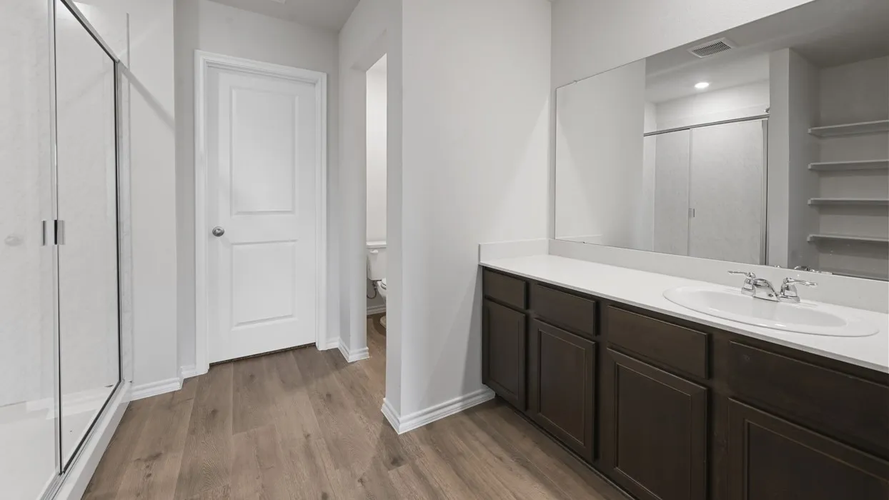 Spacious primary bathroom featuring a dark wood vanity with dual sink configuration, white quartz countertop, large framed mirror with integrated lighting, and walk-in shower with glass enclosure