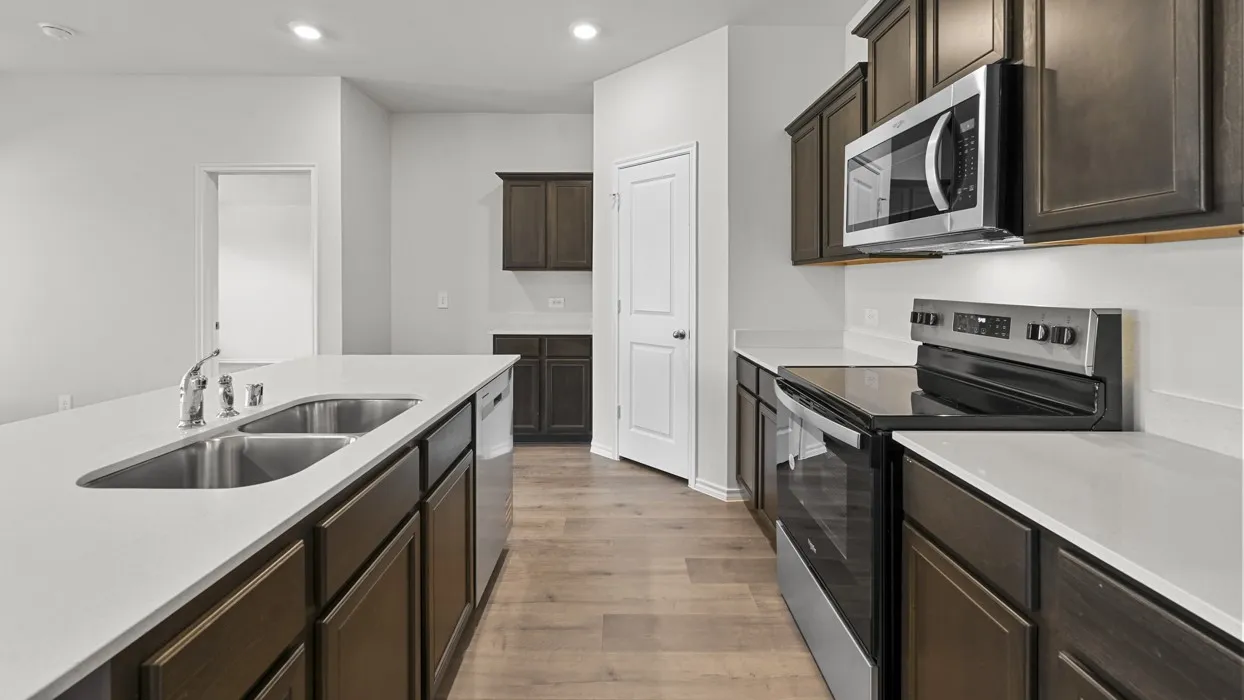 Modern galley kitchen featuring dark wood cabinetry, white quartz countertops, stainless steel appliances including a built-in microwave and electric range, a dual-basin stainless steel sink with chrome faucet, and warm wood-finish flooring throughout
