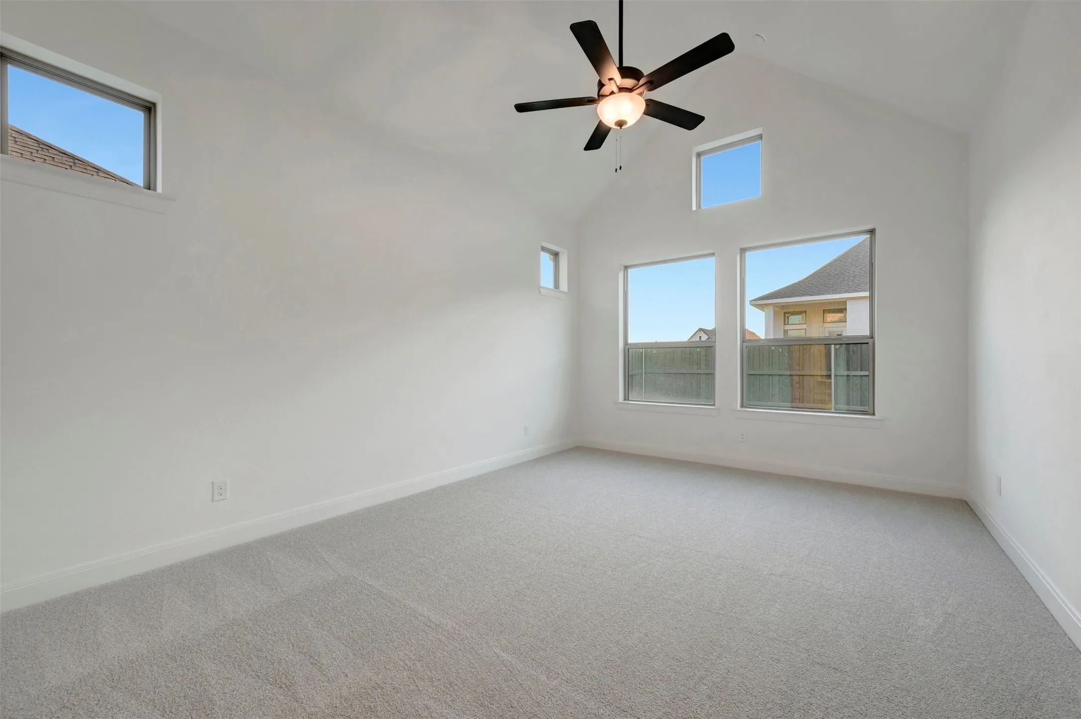 Bright, open bedroom showcasing soaring ceilings with a ceiling fan, abundant natural light from multiple windows, neutral carpet flooring, and clean white walls with minimalist finishes