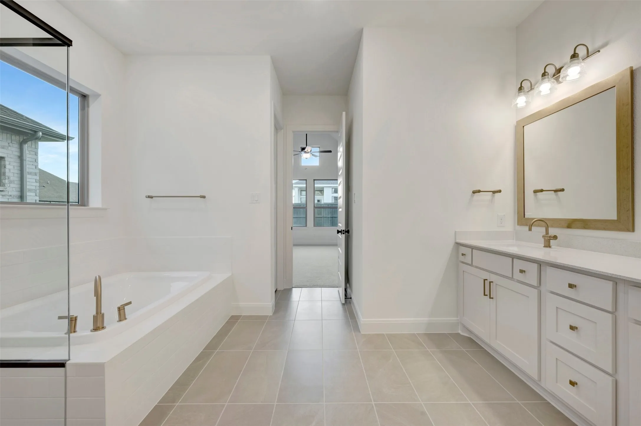 White dual vanity with gold-tone hardware, large rectangular mirror, and an adjacent built-in bathtub with gold-tone fixtures