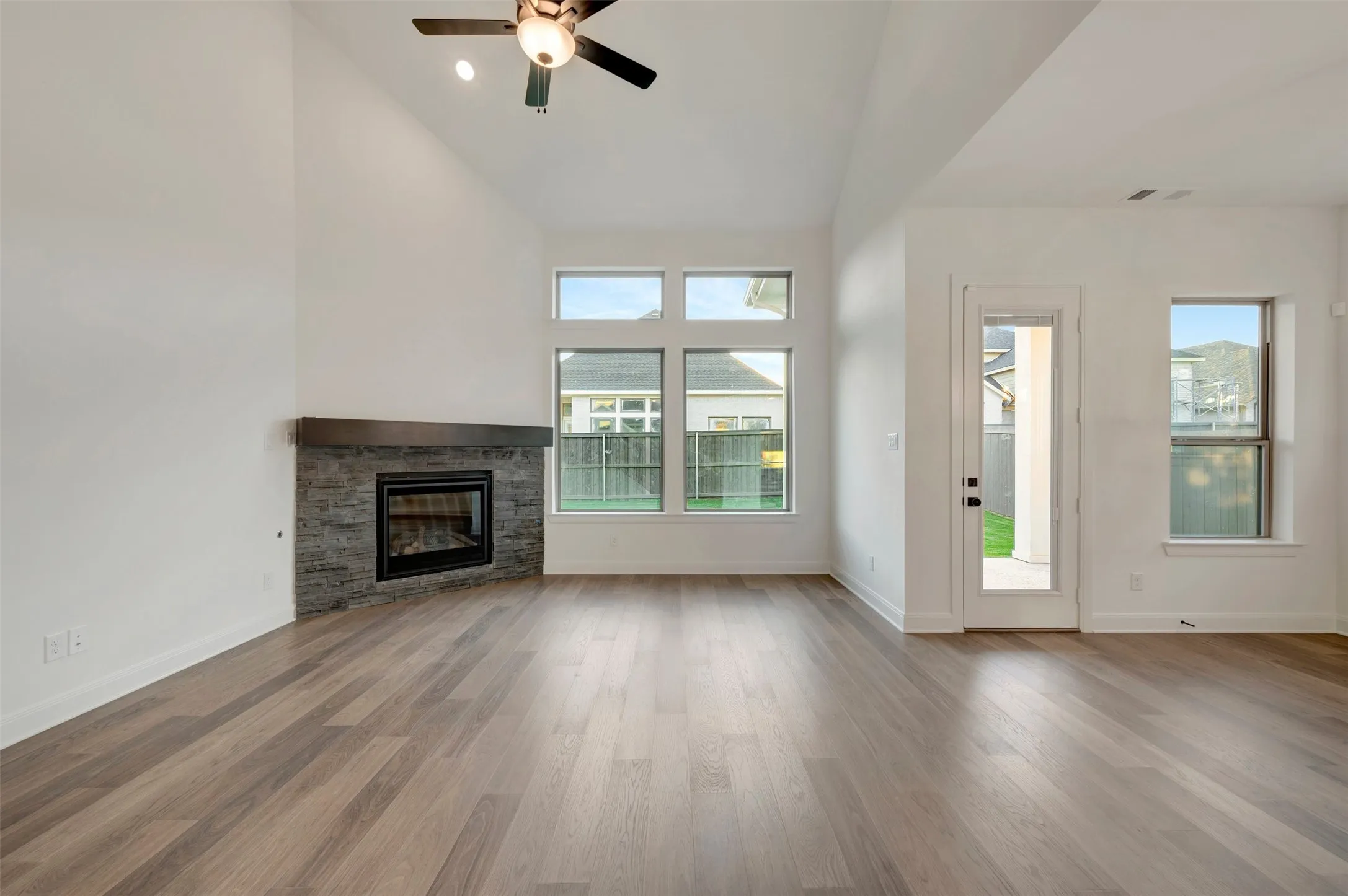 Vaulted ceiling with a ceiling fan, wood-finish flooring, fireplace with a stone surround, multiple windows, and a glass-paneled door