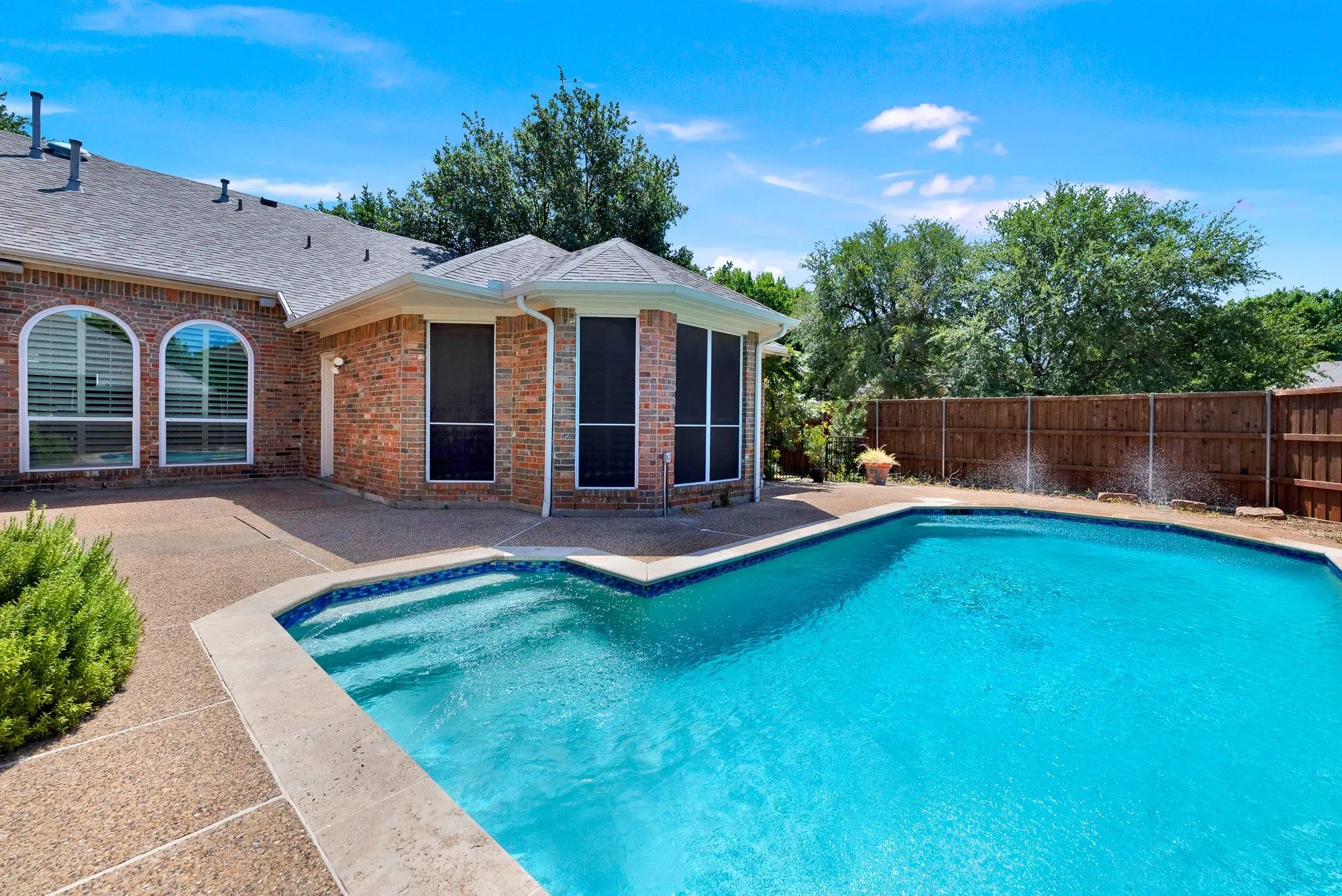 This angle showcases the built-in pool entry steps, decorative water fountains spraying into the water, and the surrounding brick home exterior. Imagine hosting sunny weekend get-togethers with friends and family enjoying the pool while you grill out on the patio.