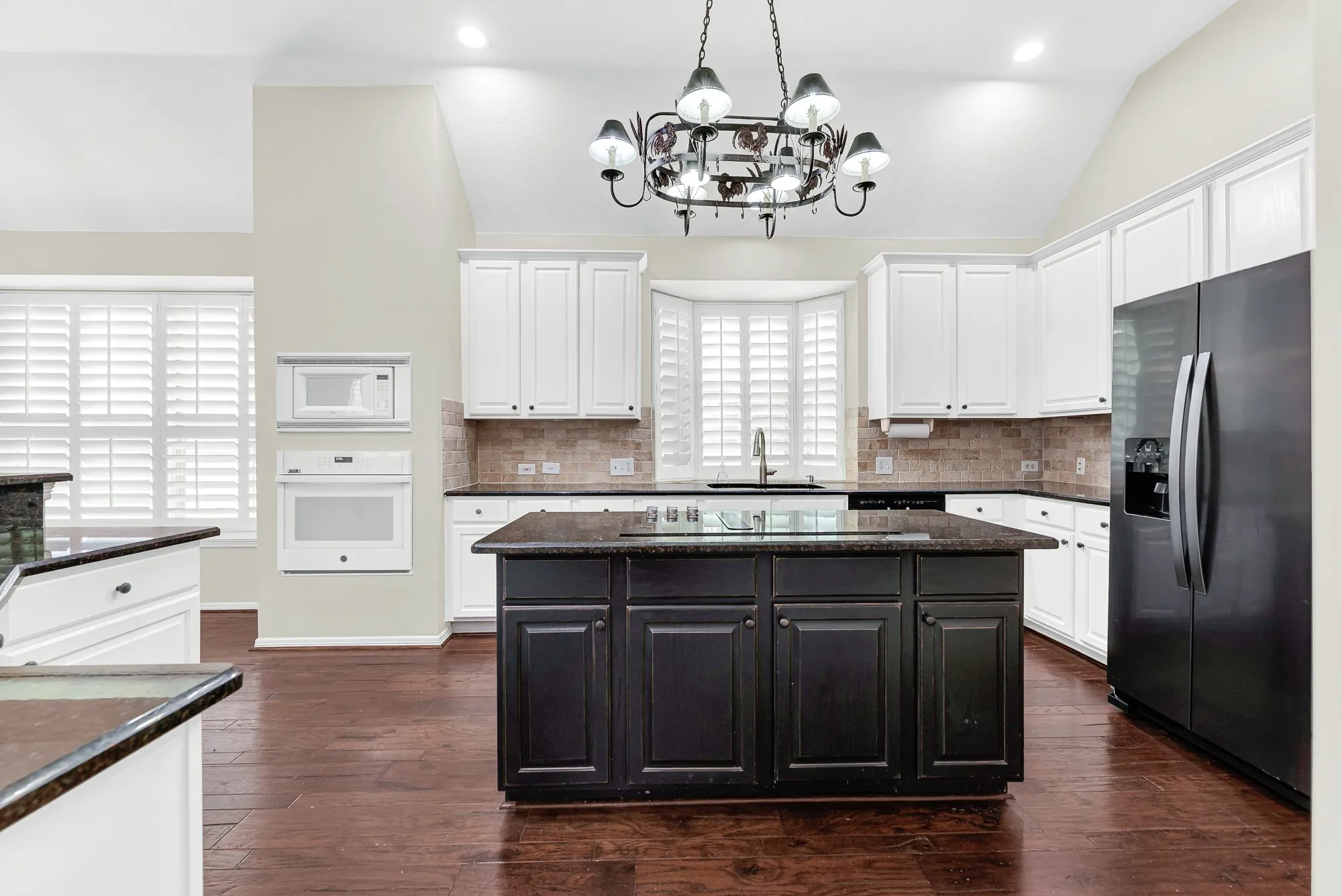 A central dark island featuring a smooth cooktop serves as the hub, framed by crisp white cabinetry, stone tile backsplash, and a statement wrought-iron chandelier. You will love cooking family meals here with everything you need right at your fingertips.