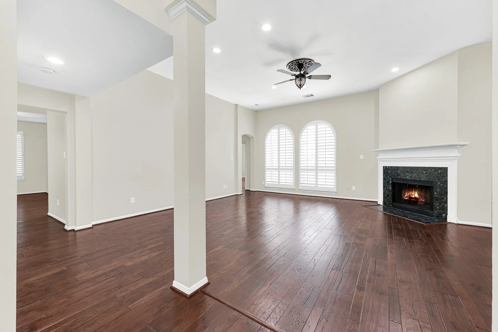 Rich hardwood flooring leads toward a cozy black-tile surround fireplace, framed by arched windows with plantation shutters and clean architectural columns. It is the ultimate spot to curl up with a cup of coffee on cool evenings or relax after a busy workday