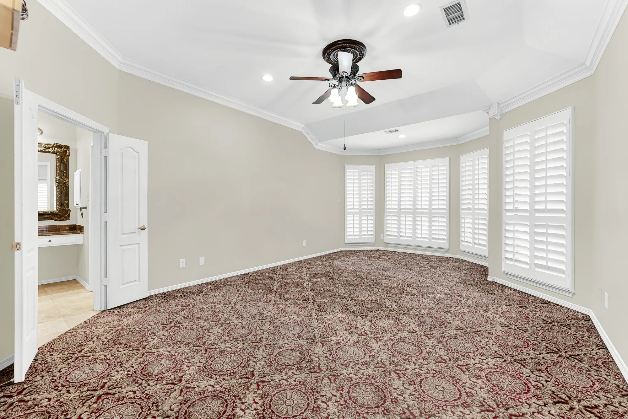 An elegant tray ceiling with crown molding caps this primary suite, featuring pattern carpeting, a ceiling fan, and direct access to the en-suite bath. It creates a serene, restful haven where you can unwind in total comfort after a long day.