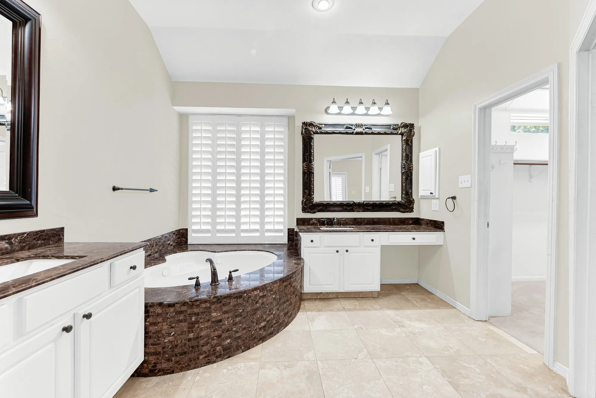Looking across the tiled floor reveals a dedicated vanity seating area with a large carved mirror, plantation-shuttered window, and a door into the walk-in closet. It creates a convenient, organized environment that makes getting ready for morning routines or evening events effortless.