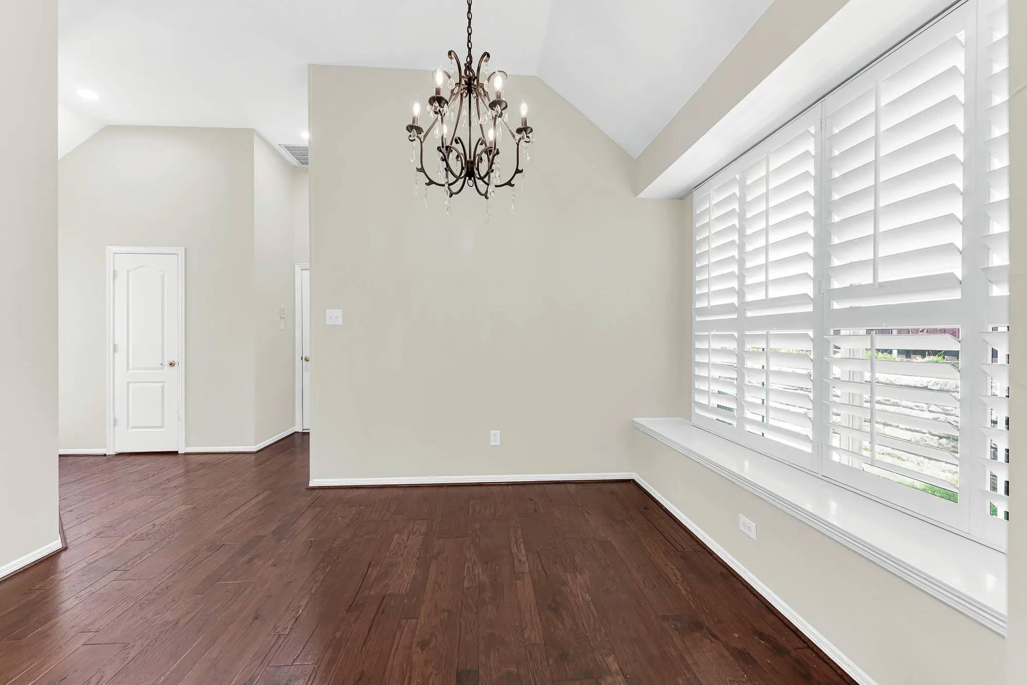 Sunlight pours through wide plantation shutters onto a deep window bench and rich hardwood floors under a delicate chandelier. This charming nook is perfect for pulling up a table for casual morning breakfasts or relaxing with weekend coffee.