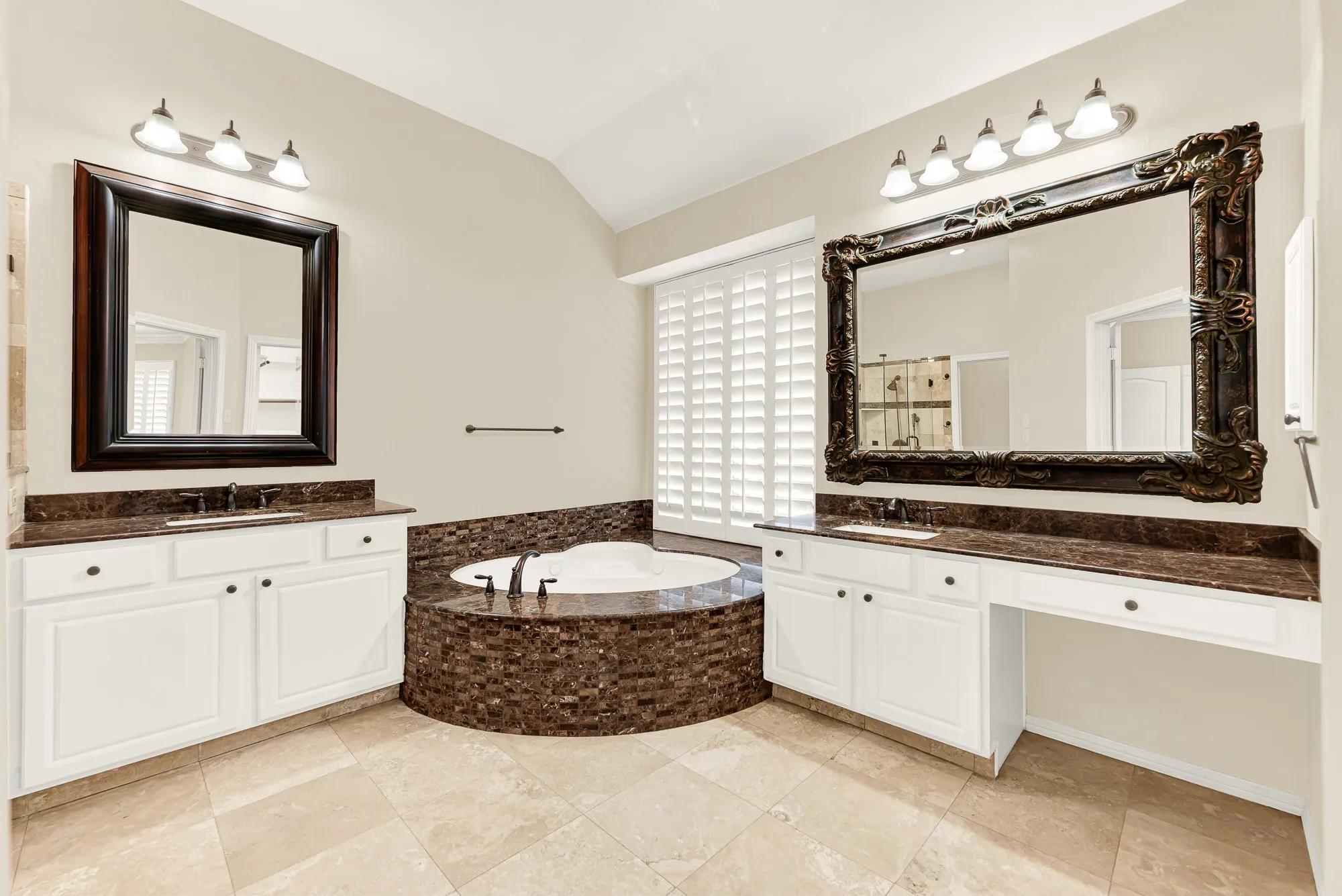 A luxurious corner soaking tub wrapped in rich mosaic stone serves as the centerpiece, flanked by dual white vanities with dark granite tops. It offers a spa-like retreat where you can soak away the day in total calm and luxury.