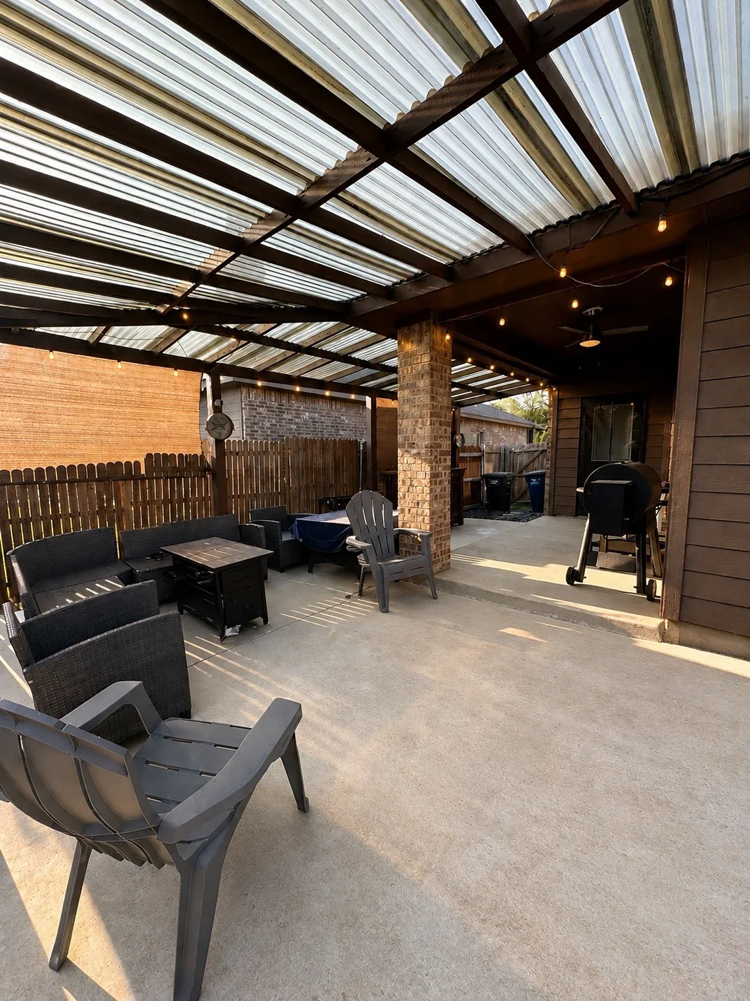 Expansive outdoor patio with a corrugated roof, brick support columns, string lighting, and wood plank siding