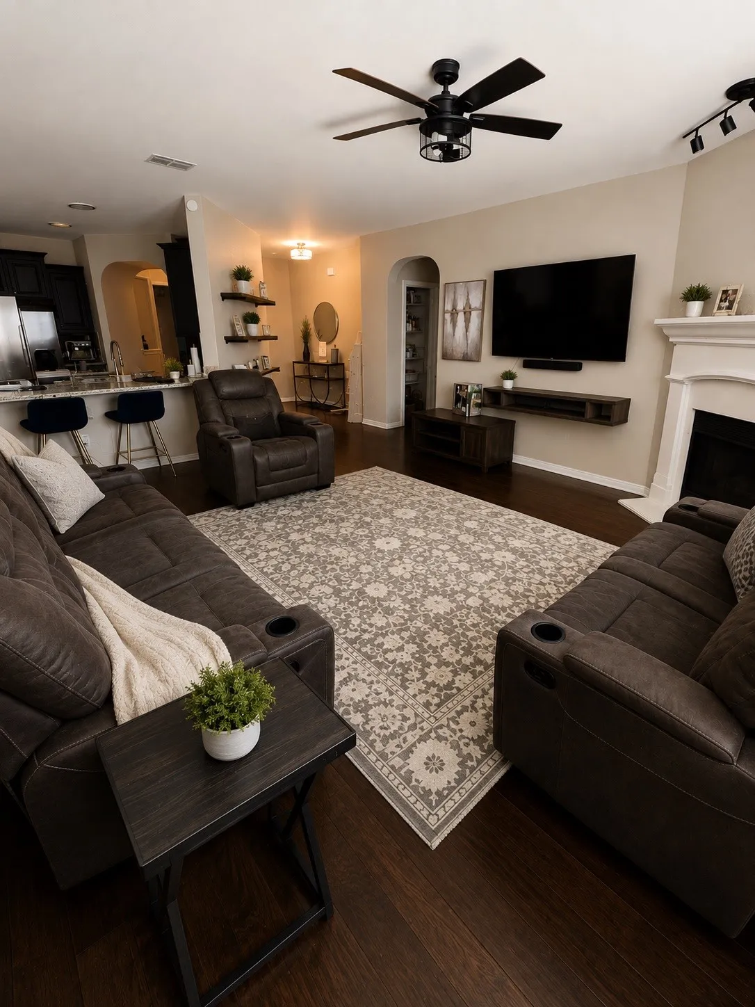 Wood-finish flooring throughout, featuring a kitchen island with seating, built-in shelving, fireplace with mantel, and a ceiling fan with integrated lighting