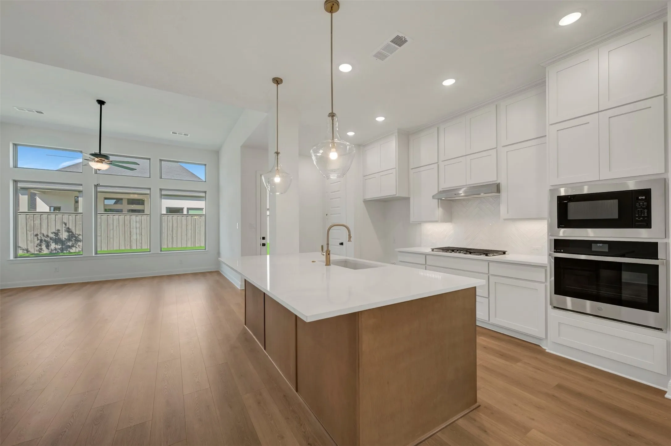 Open-concept living space featuring wood-finish flooring, a contemporary kitchen island with pendant lighting, white shaker cabinetry, stainless steel appliances, and a wall of windows