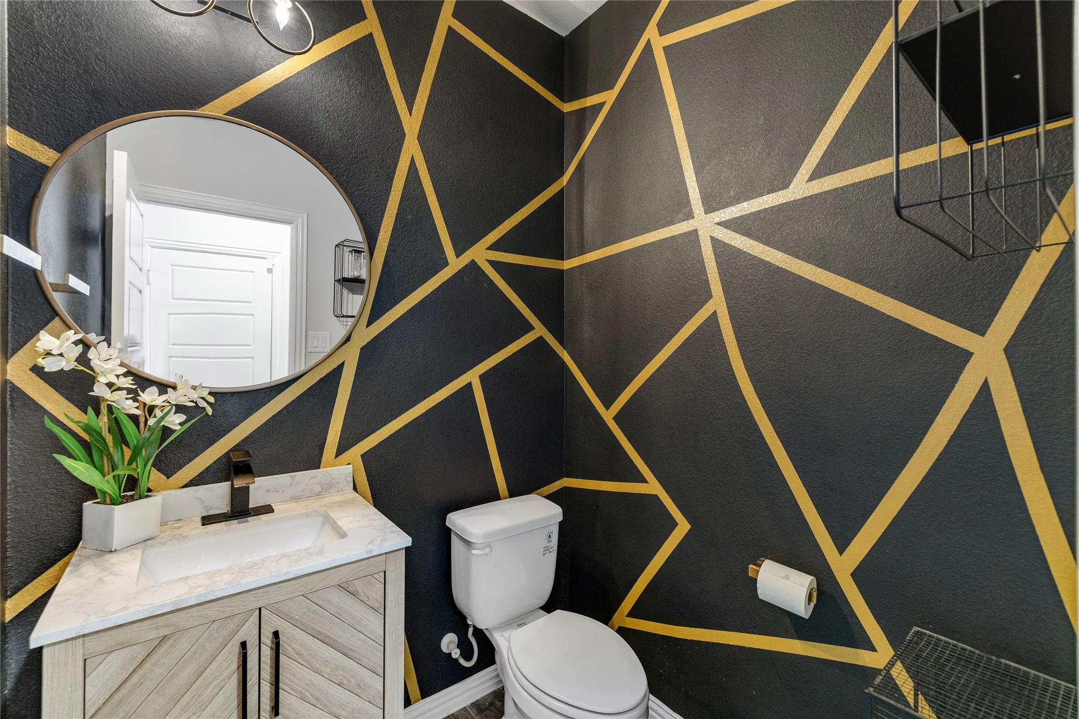 Geometric accent wall, modern vanity with wood-finish doors, integrated sink, and an oval mirror with a metal frame