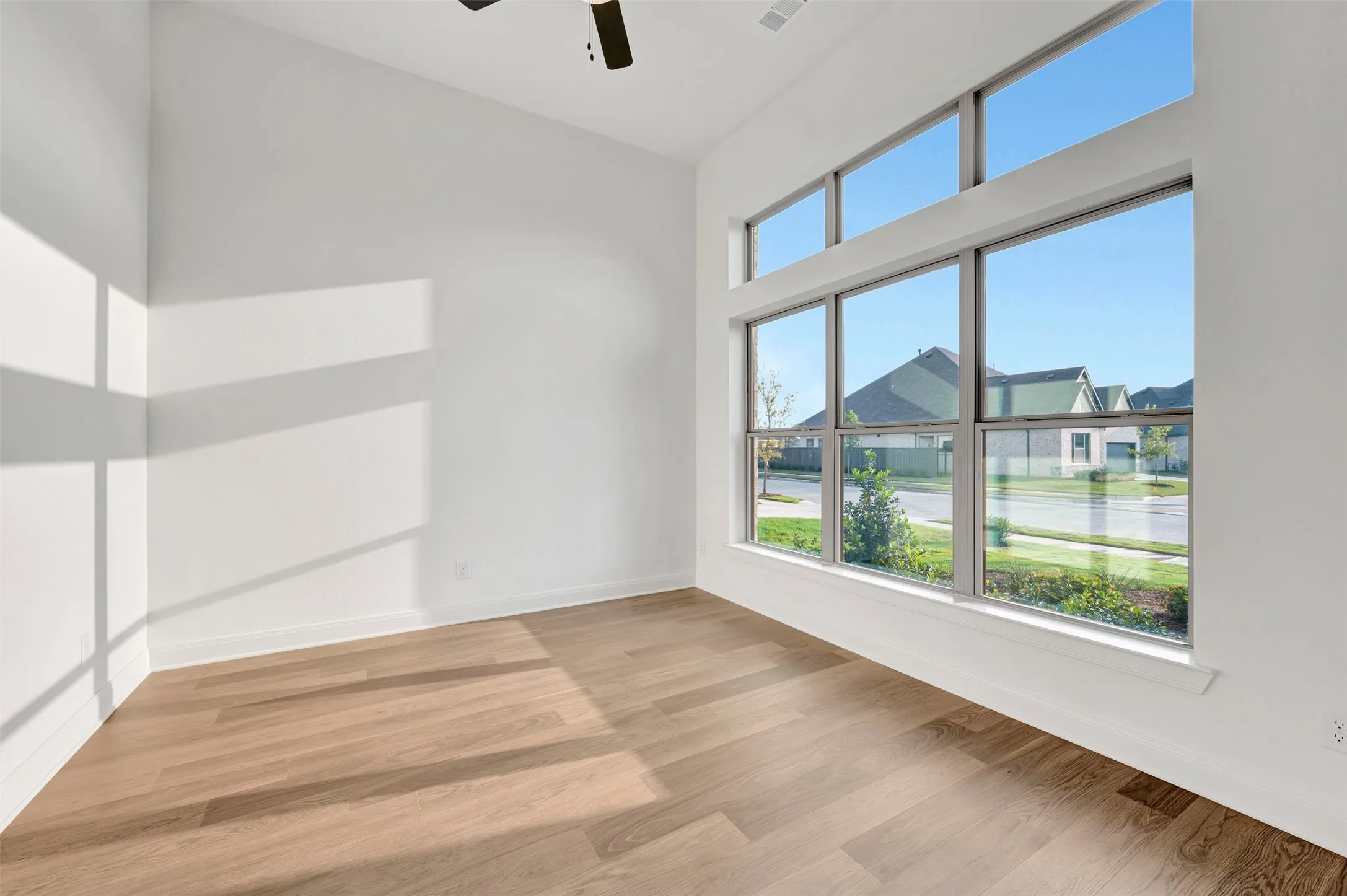 Bright corner room showcasing expansive multi-pane windows framing neighborhood scenery, warm wood-finish flooring, white walls, and abundant natural light throughout the space
